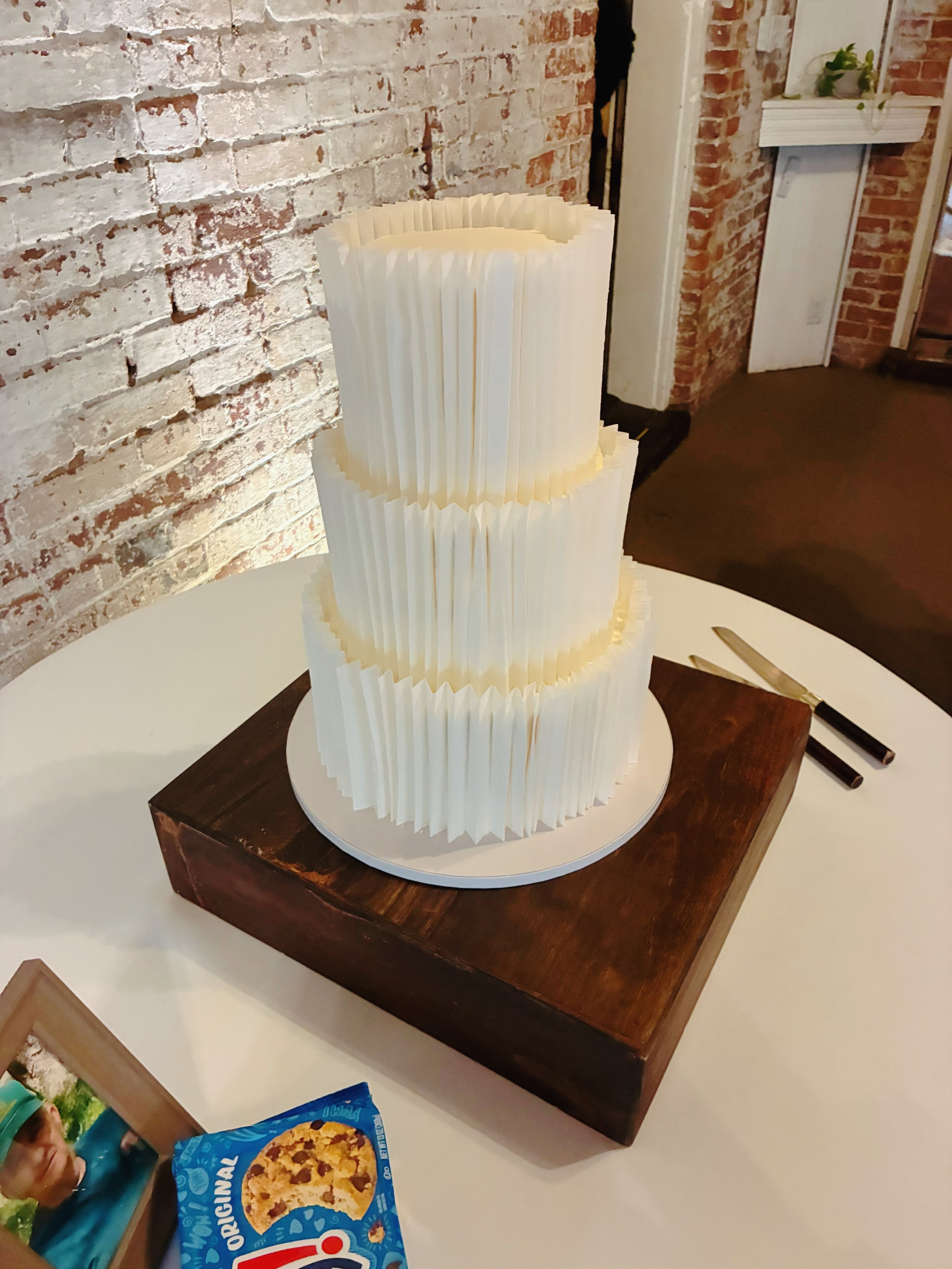 A paper sculpture resembling a cupcake or layered dessert on a white base, placed on a dark wooden table against an exposed brick wall with a white wooden trim and some houseplants.