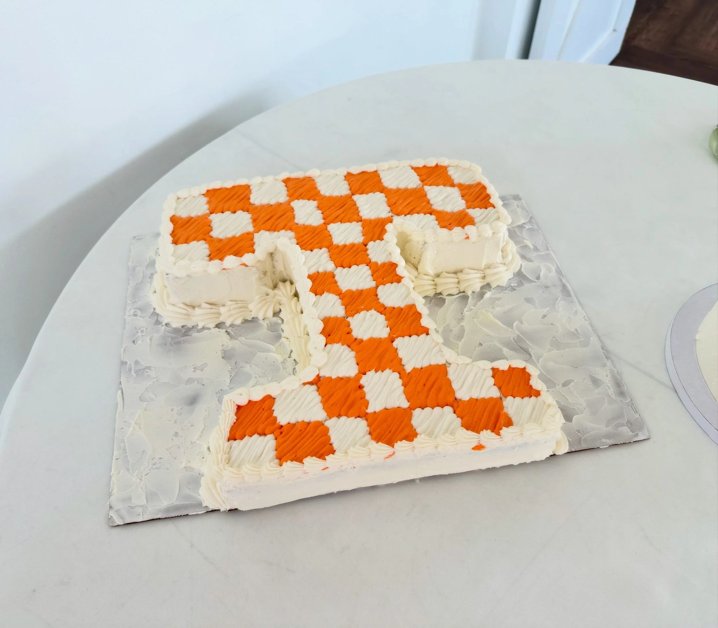 A rectangular white frosted cake with orange and white checkered pattern on top, partially cut, placed on a white surface.