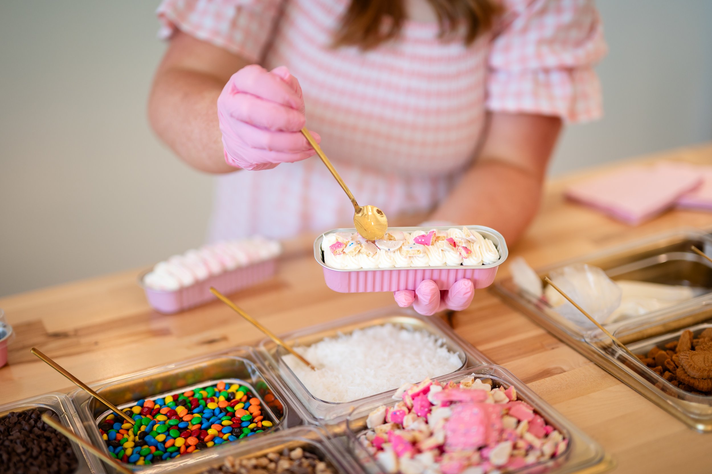 A person wearing pink gloves is using a golden spoon to scoop pink frosting from a container, surrounded by various dessert toppings like sprinkles, candies, and cookies.