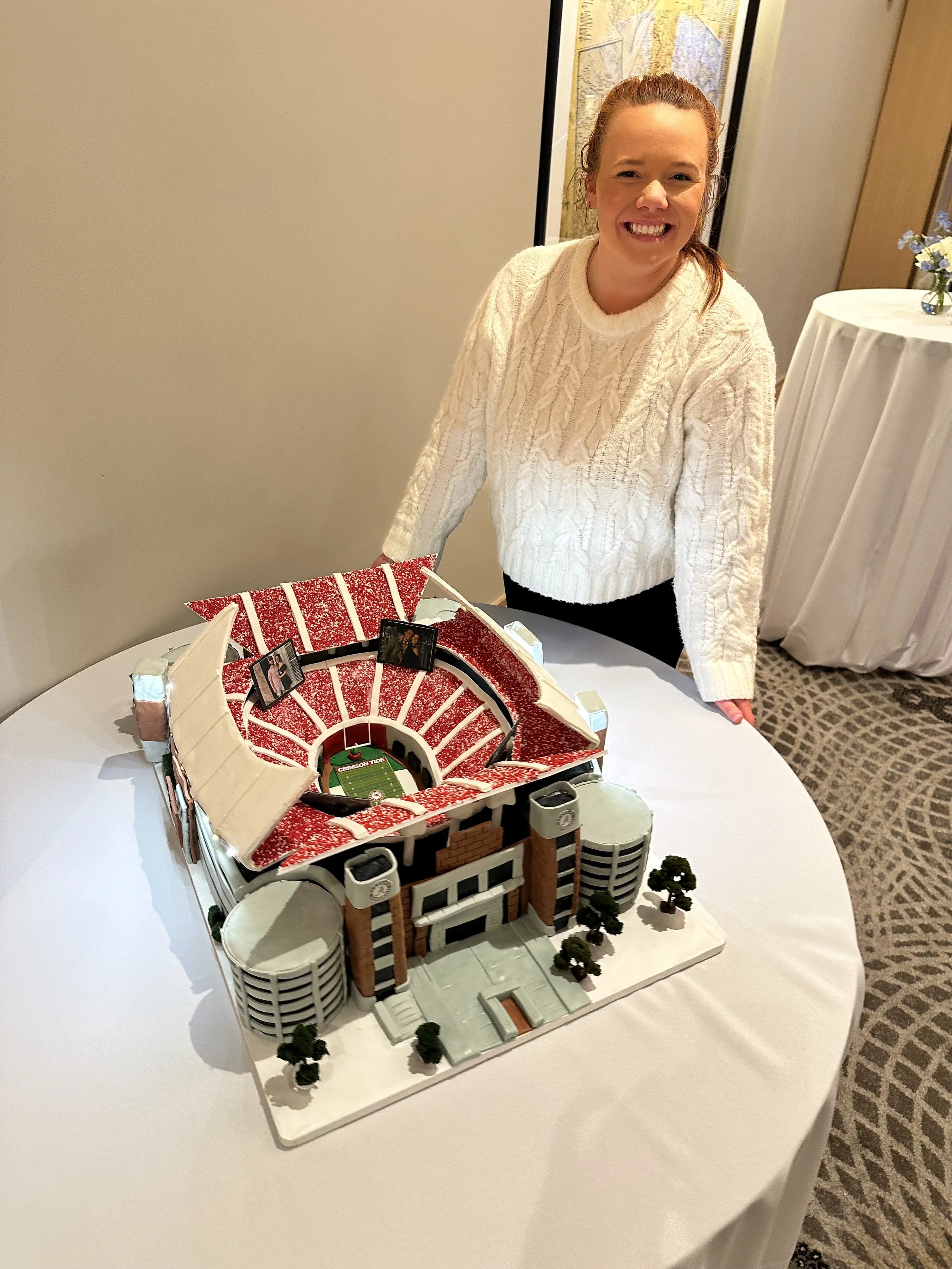 A woman is smiling while standing behind a scale model of a stadium on a round table. The model features red seats, small trees, and miniature details. She is wearing a cream-colored cable-knit sweater, and part of a framed map is visible on the wall