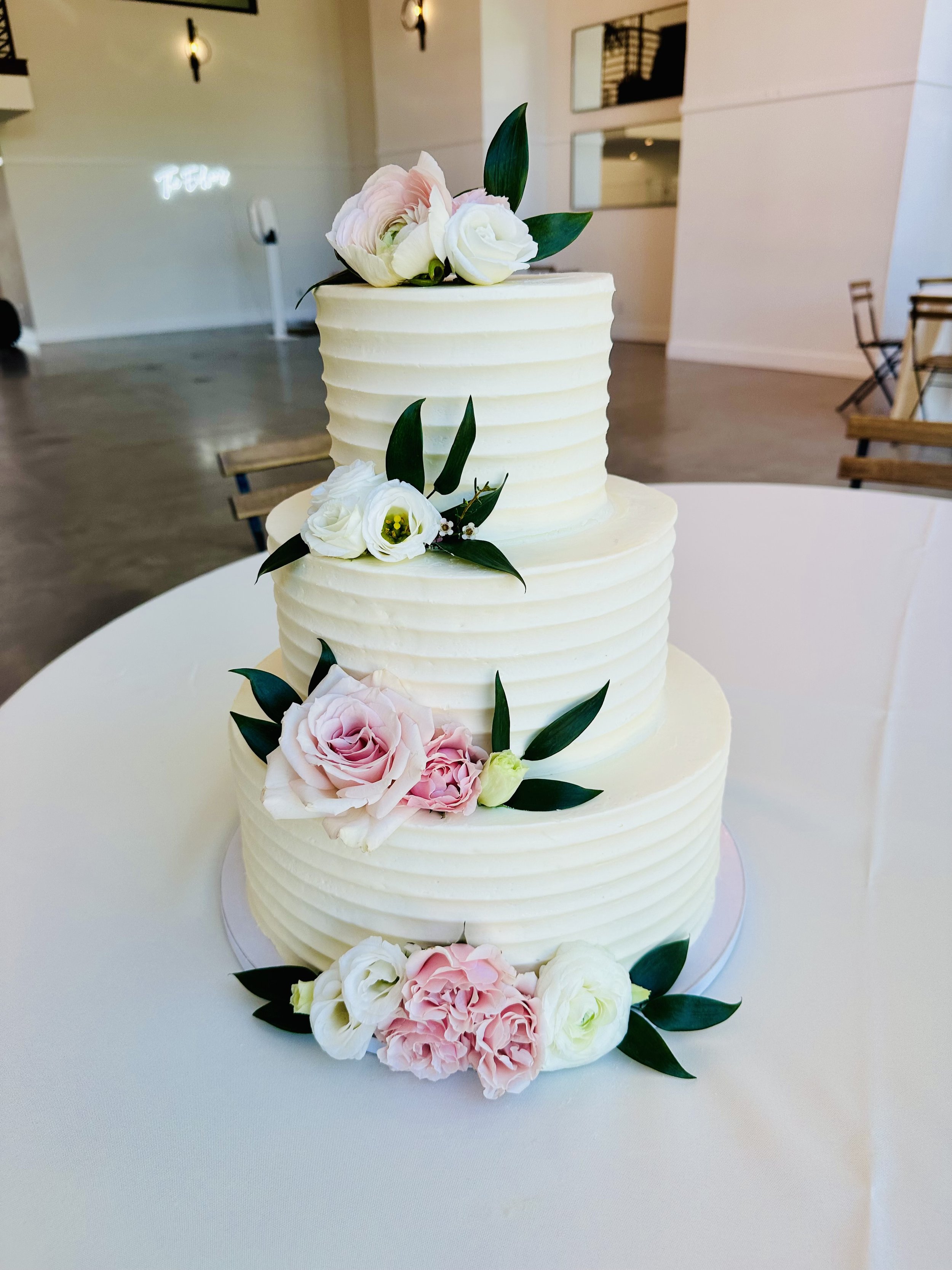 A three-tiered white wedding cake decorated with fresh pink and white roses and green leaves, set on a white table in a modern indoor setting.