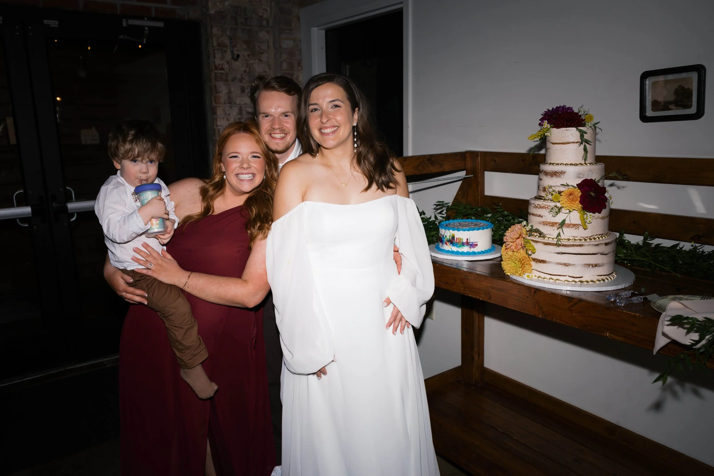 A group of four people, including a woman in a white dress, a woman in a maroon dress, a man, and a young boy, smiling and posing at a celebration with a decorated cake on a wooden table.