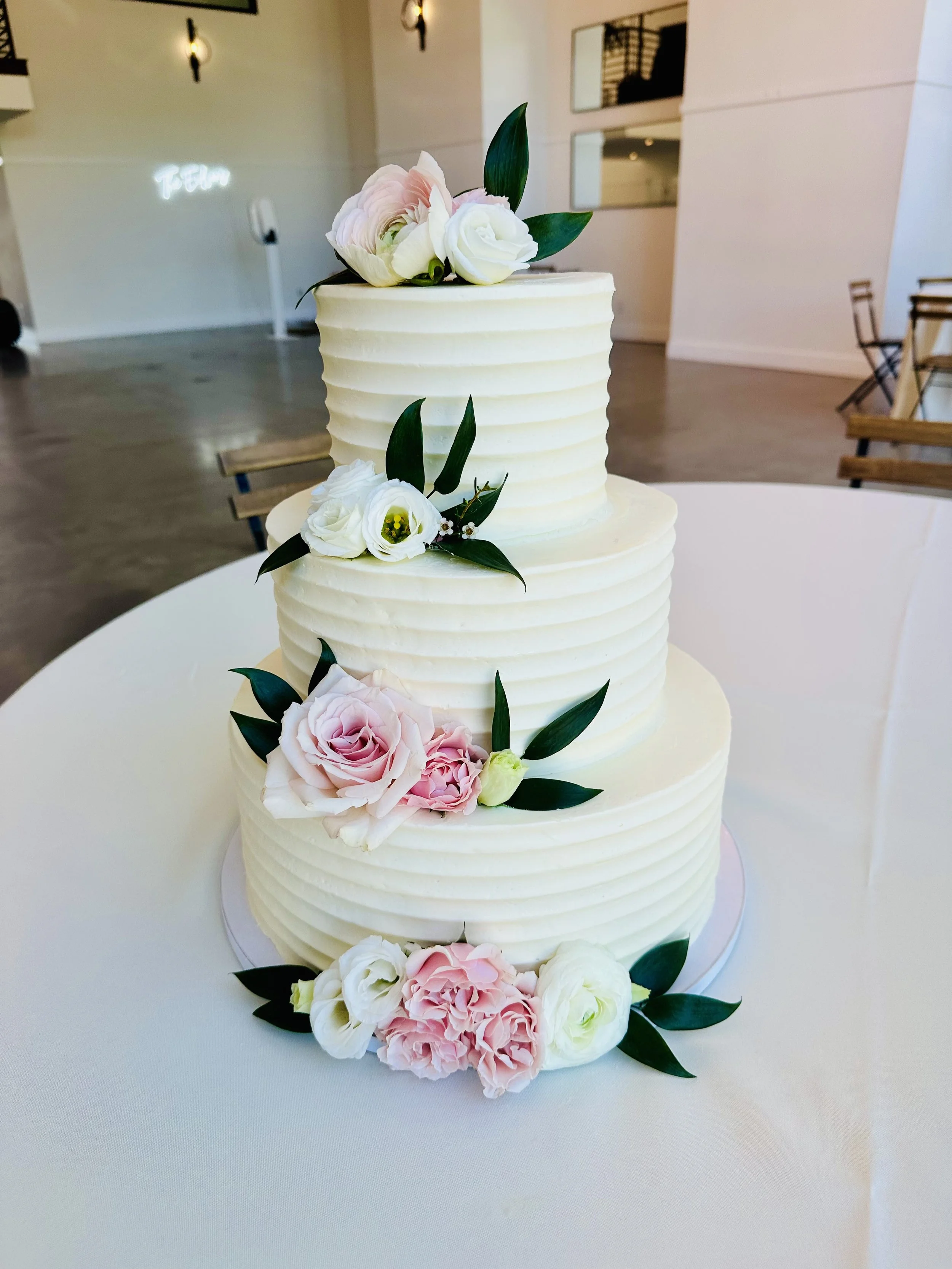 A three-tier white wedding cake decorated with pink and white flowers and green leaves, placed on a white table in a modern interior.