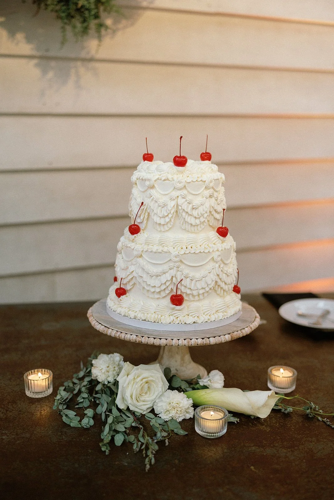 Three-tiered white wedding cake decorated with whipped cream swirls and topped with maraschino cherries, placed on a cake stand with white flowers and candles surrounding it.