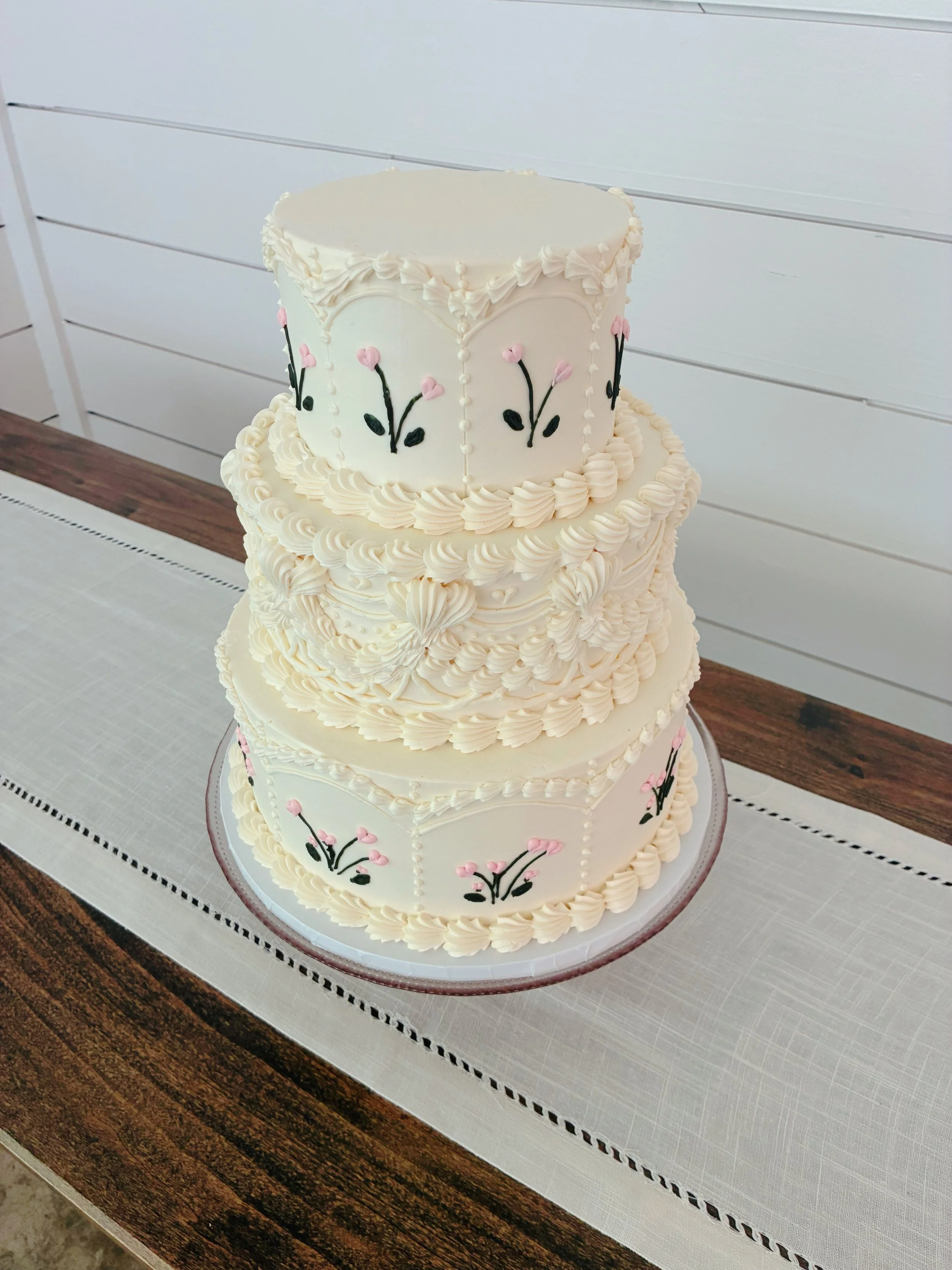A three-tier white wedding cake decorated with pink flowers and black vines, sitting on a white cake board with a pink ribbon border, on a wooden table with a gray table runner.
