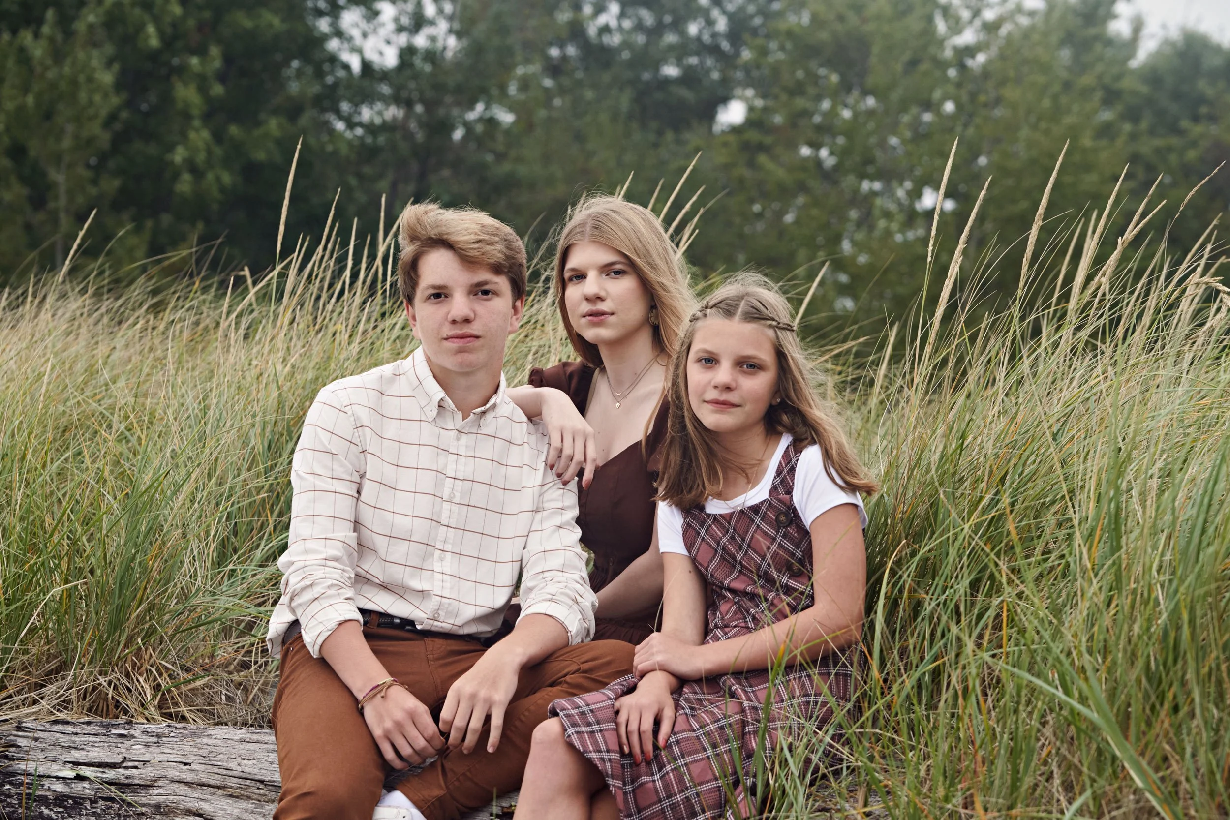 Family portrait of three young siblings sitting on a log in a grassy outdoor setting.