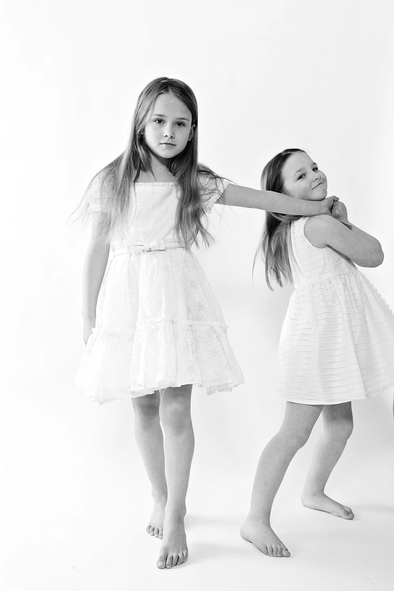 two little sisters in white dresses. the younger one is pulling the older one away. black and white image on white background. portsmouth NH