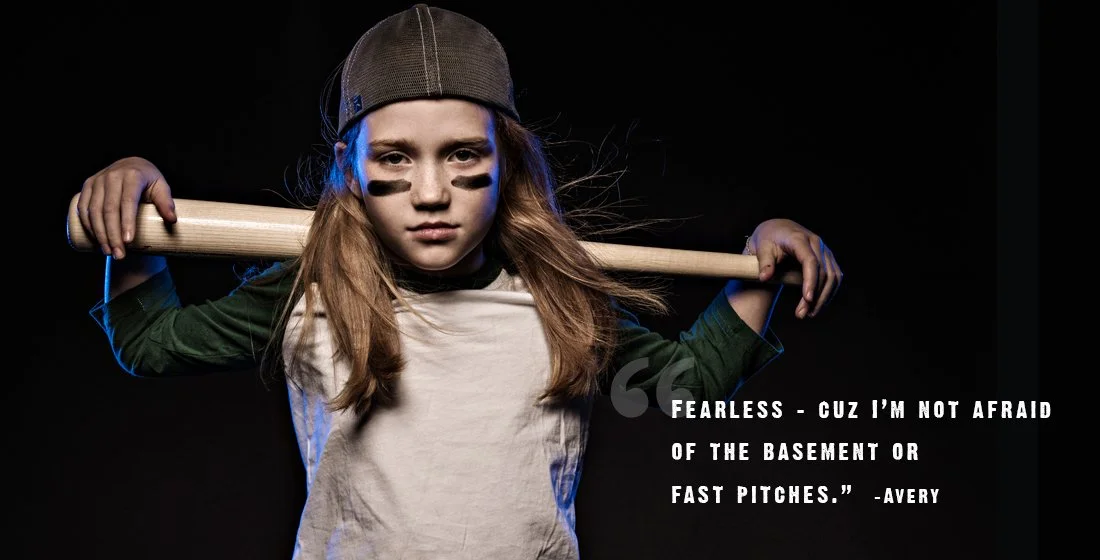 little girl with base ball bat over her shoulders, hat on backwards, with eye black under her eyes, against a black background. gritty. tough. Portsmouth NH