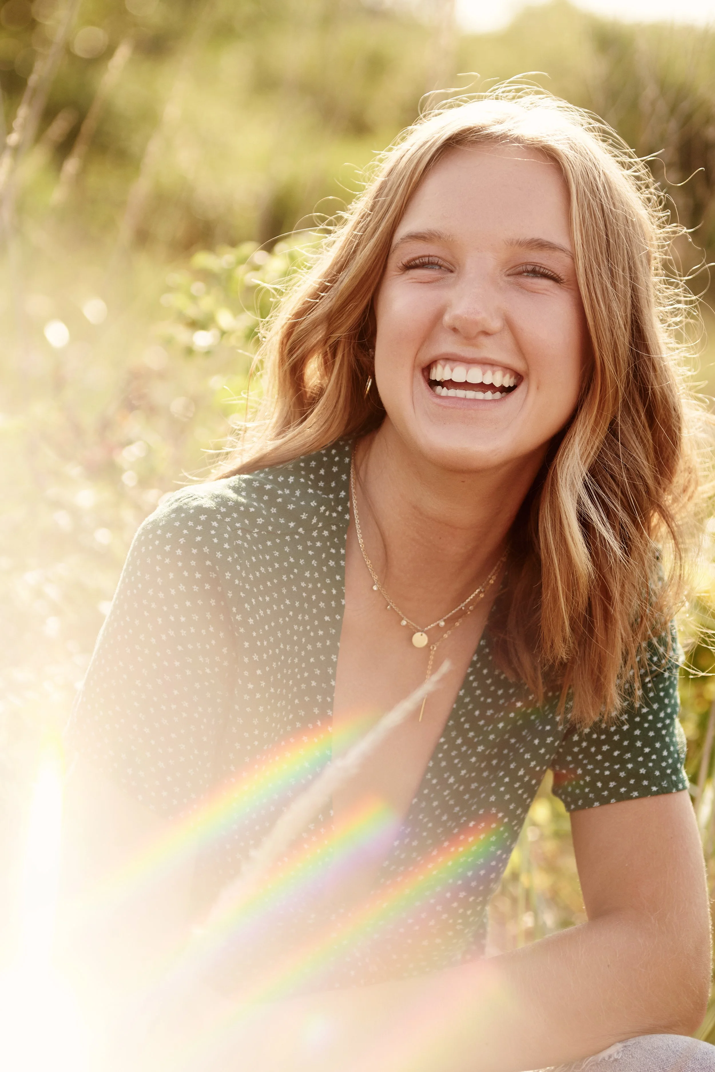 Senior portrait of young woman smiling sitting outdoors with sunlight and rainbow lens flare.