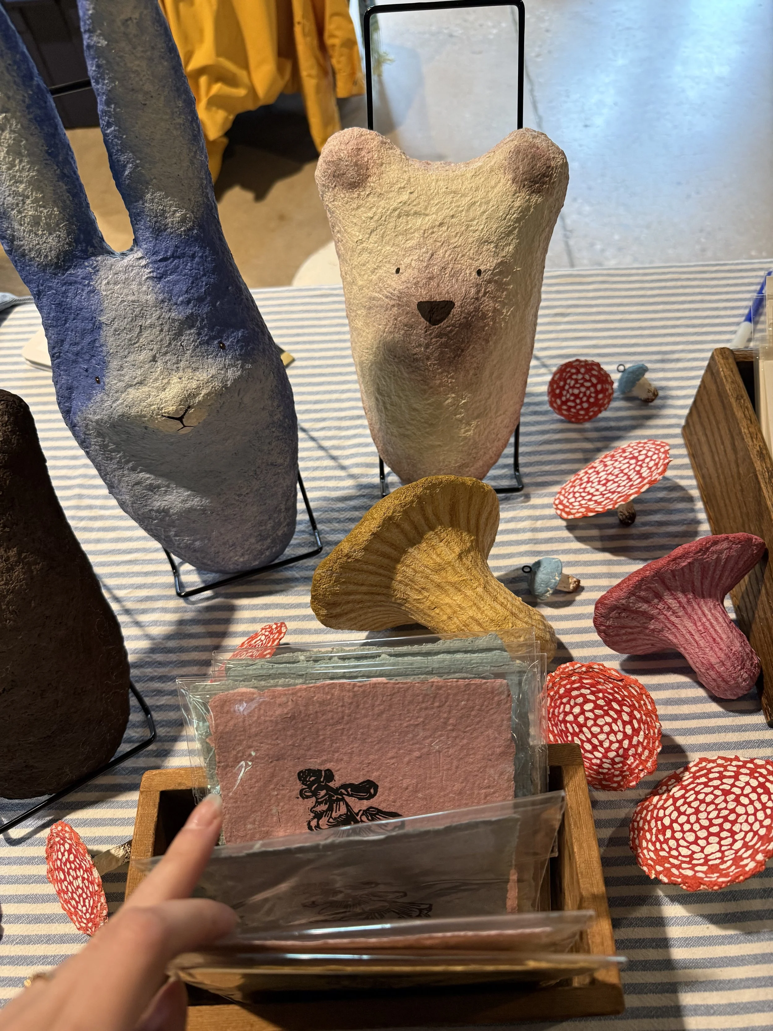 Close-up of a handmade art market display featuring a cream-colored paper-clay bear and blue rabbit sculpture, surrounded by colorful paper clay mushrooms and small handmade decorations. A wooden box holds colorful handmade paper and prints