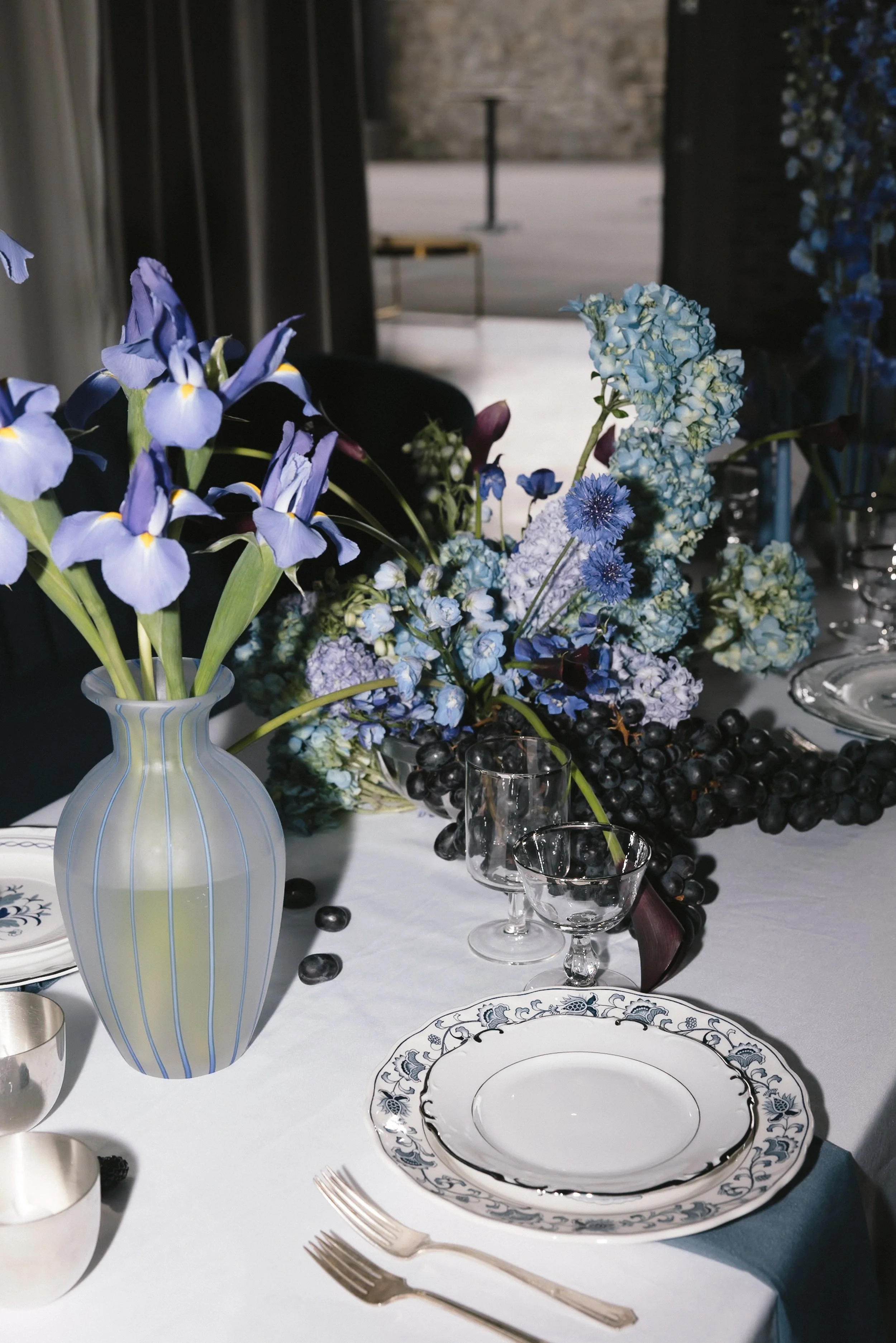 An elegant table setting with blue and white floral centerpiece, including irises, hydrangeas, and other flowers, on a white tablecloth with patterned plates, silverware, and glassware.