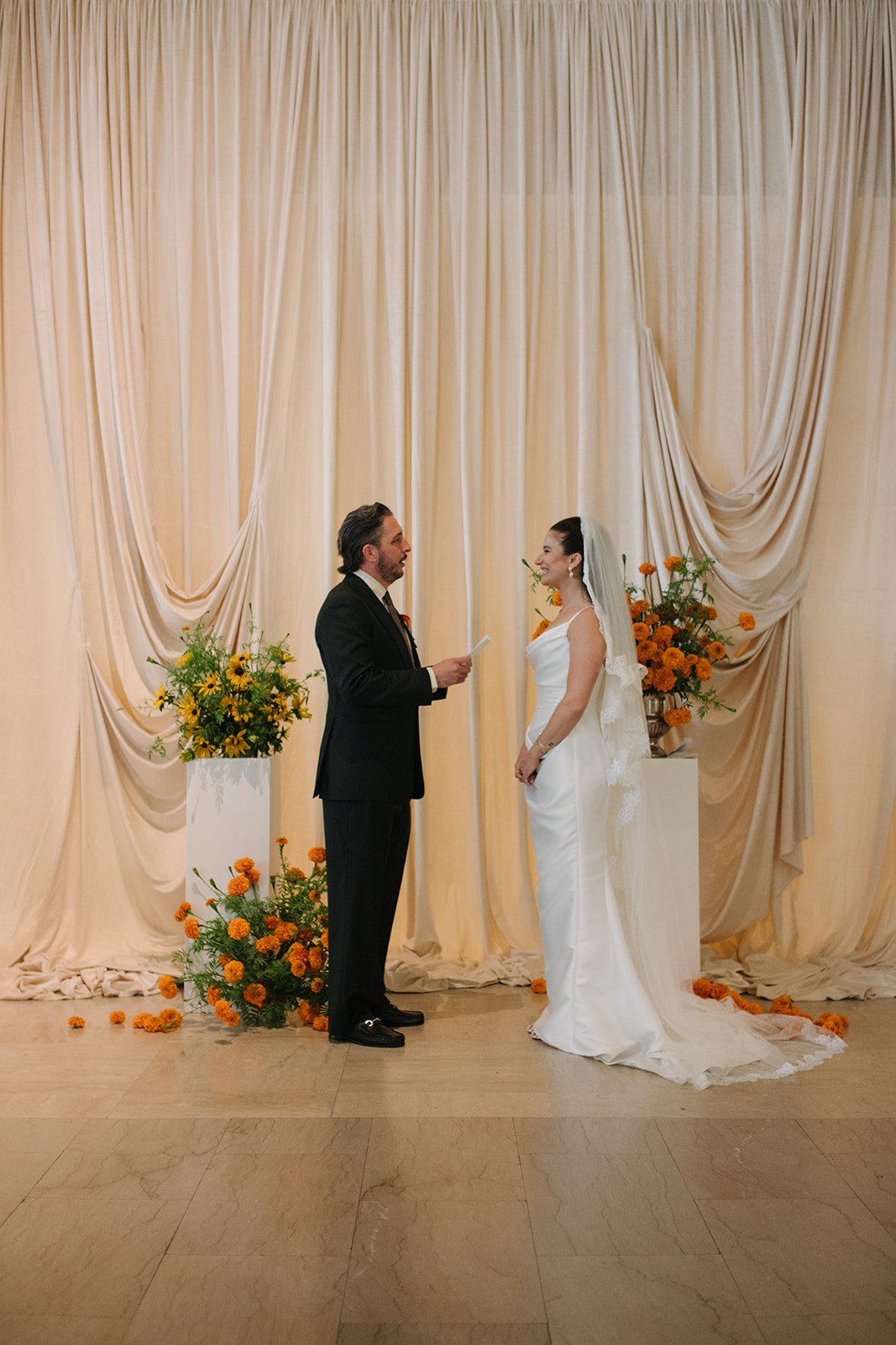 Modern september wedding ceremony ivory draped fabric orange and yellow color palette floral pedestals marigolds library wedding ceremony bride and groom  the cottage rose kansas city wedding florist crossroads hotel kansas city wedding