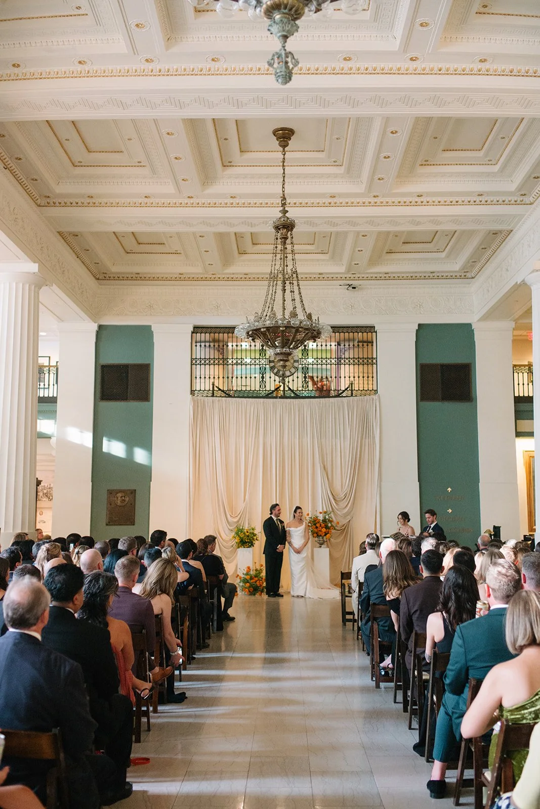 Modern september wedding ceremony ivory draped fabric orange and yellow color palette floral pedestals marigolds library wedding ceremony bride and groom  the cottage rose kansas city wedding florist crossroads hotel kansas city wedding
