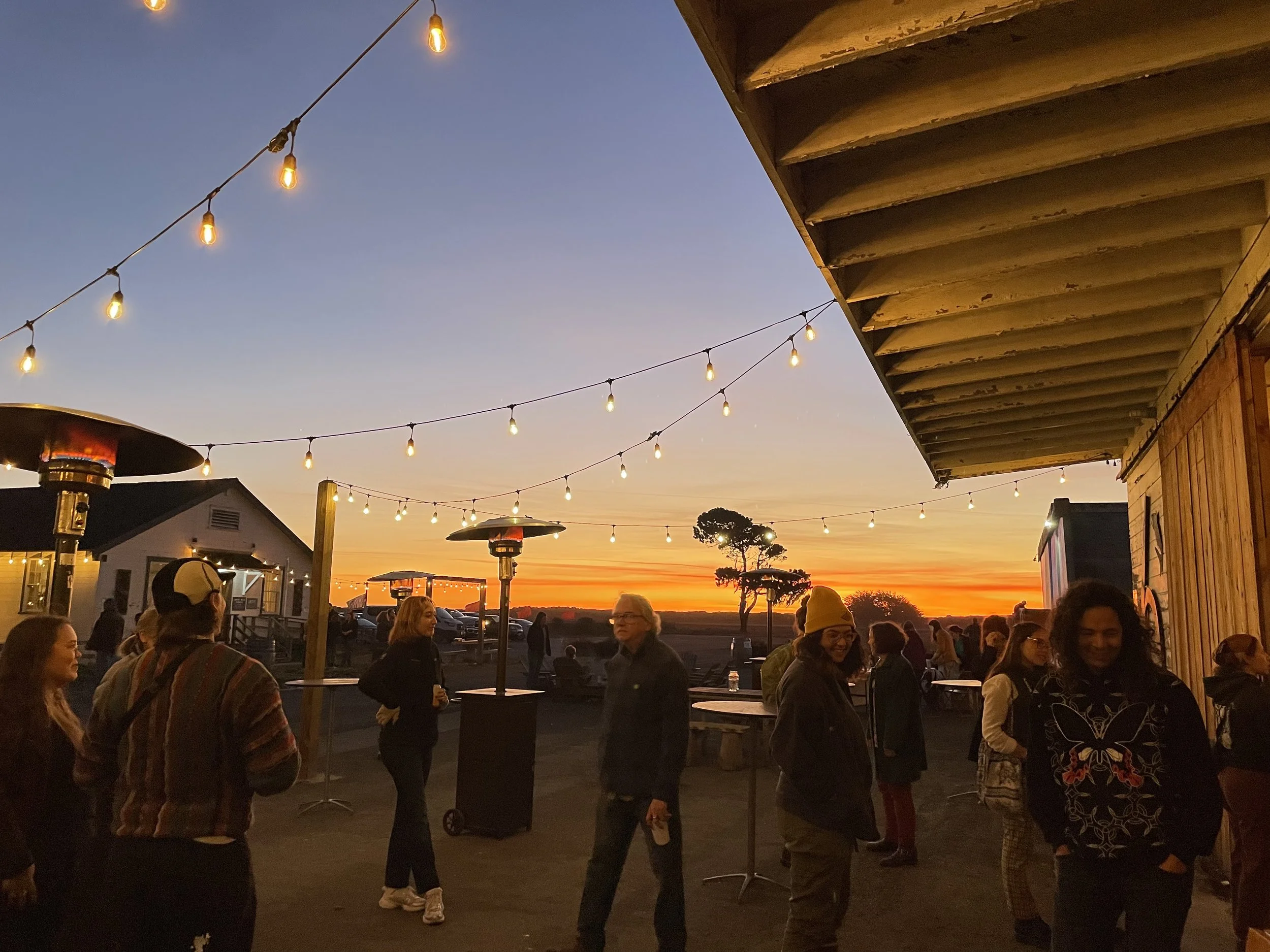 Outdoor gathering at sunset with string lights and people socializing. Patio heaters are present, and a building structure is visible. The sky is a gradient of blue and orange hues.