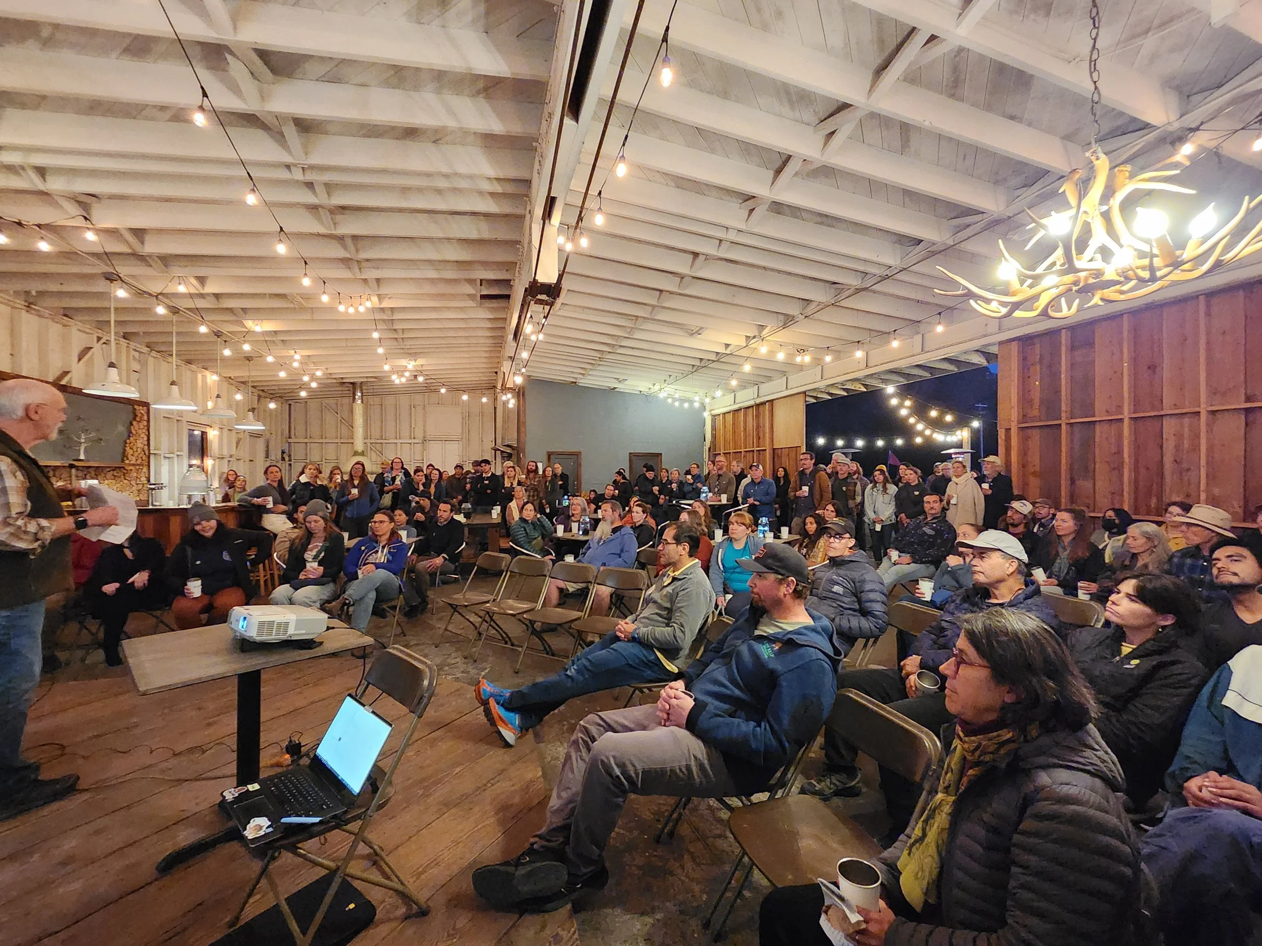 A speaker addressing a large audience seated in a rustic venue with wooden walls and string lights. The audience members are attentive, some sitting with drinks, others standing. The setting includes a projector, laptop, and decorative ceiling lights