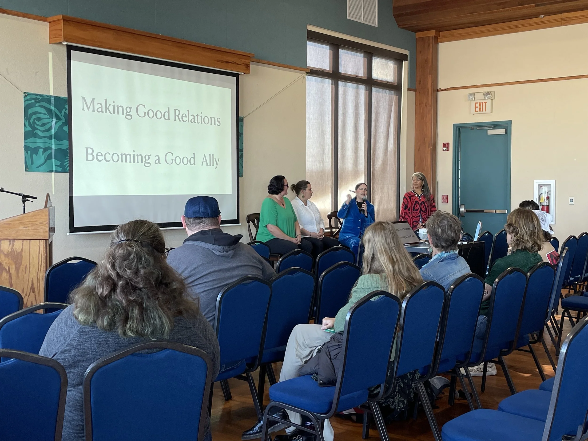 A group seminar with attendees seated in blue chairs facing a panel of speakers. A projection screen displays the text 'Making Good Relations, Becoming a Good Ally.' The room has a wooden floor and a podium on the left.