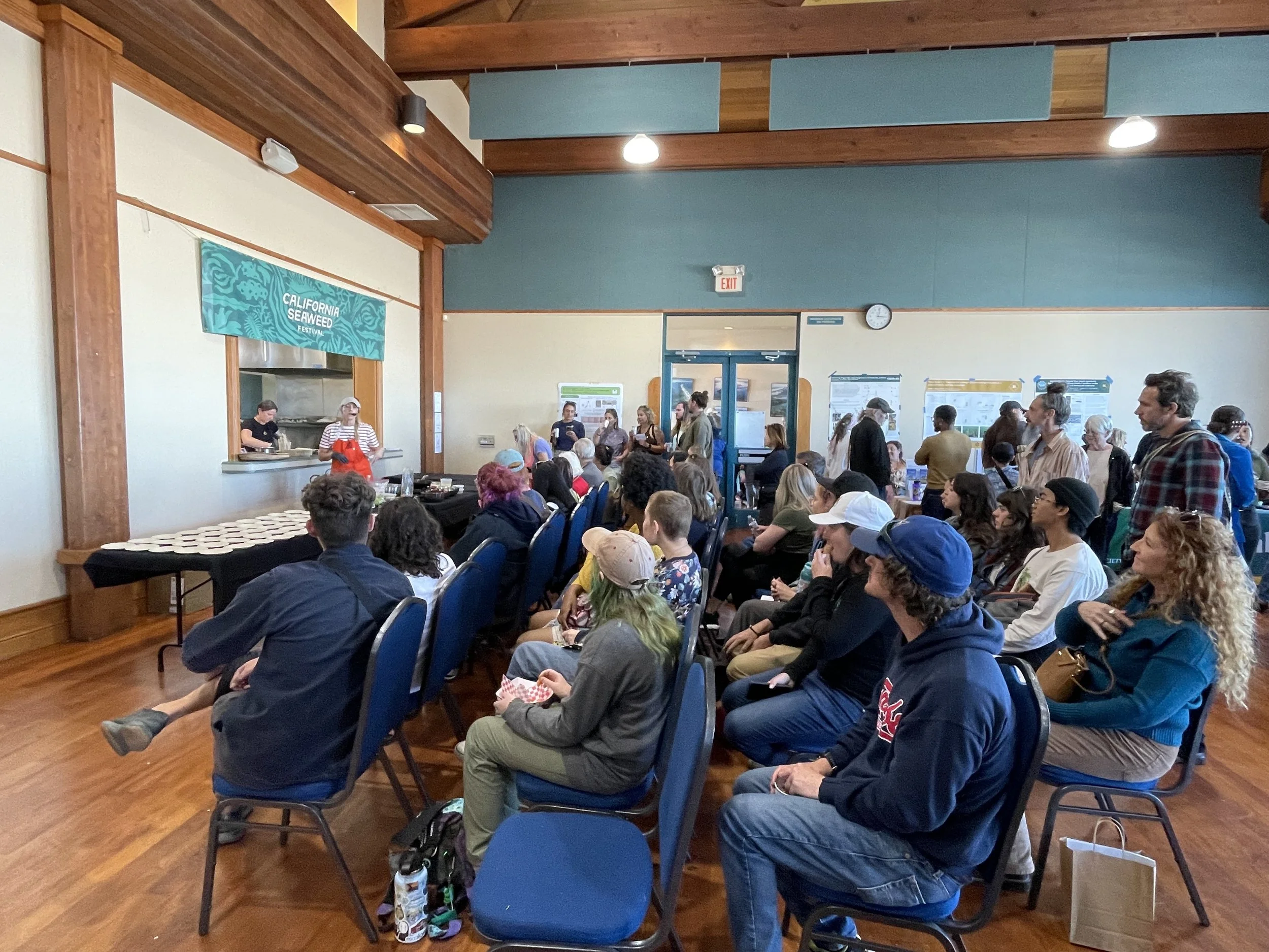 People attending a cooking demonstration at the California Seaweed Festival, with a chef presenting at a table with plates, and an audience seated and standing.