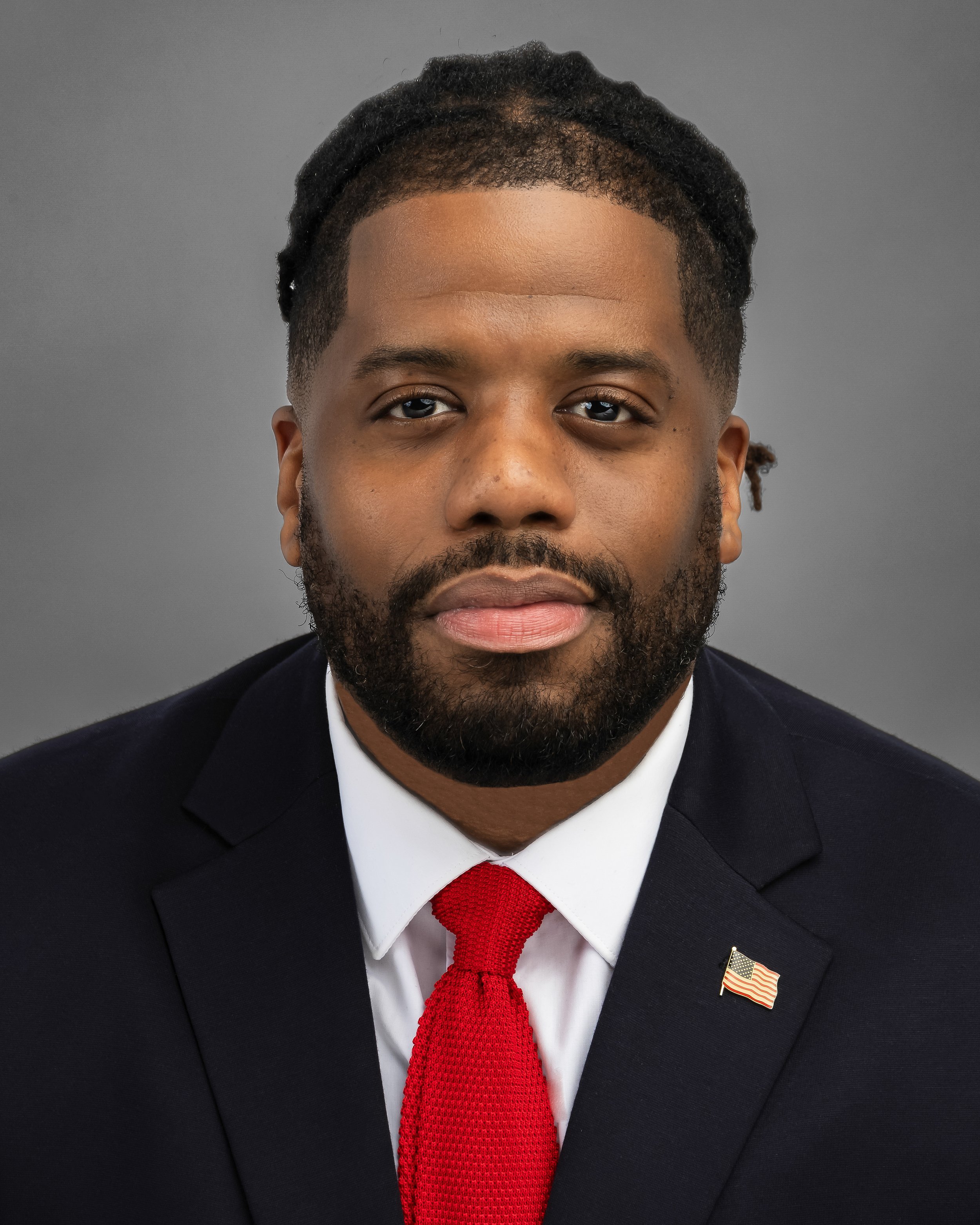 Man wearing a dark suit with an American flag pin, smiling against a gray background.