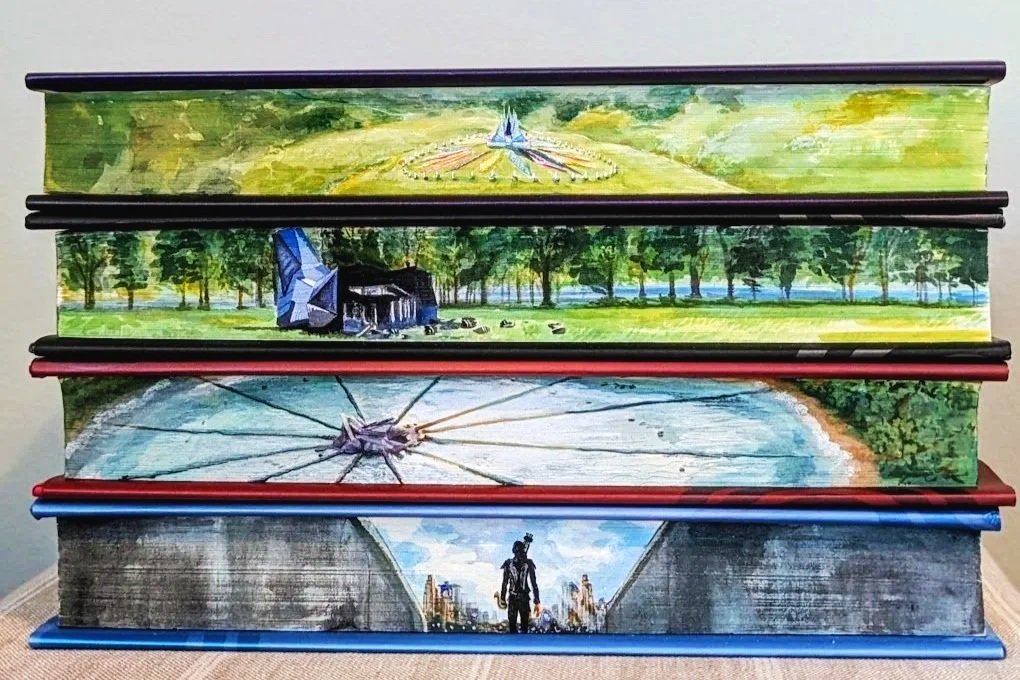 Stacked books with painted covers depicting a futuristic landscape, an airship, a lake with a large structure and a person walking towards a city skyline.