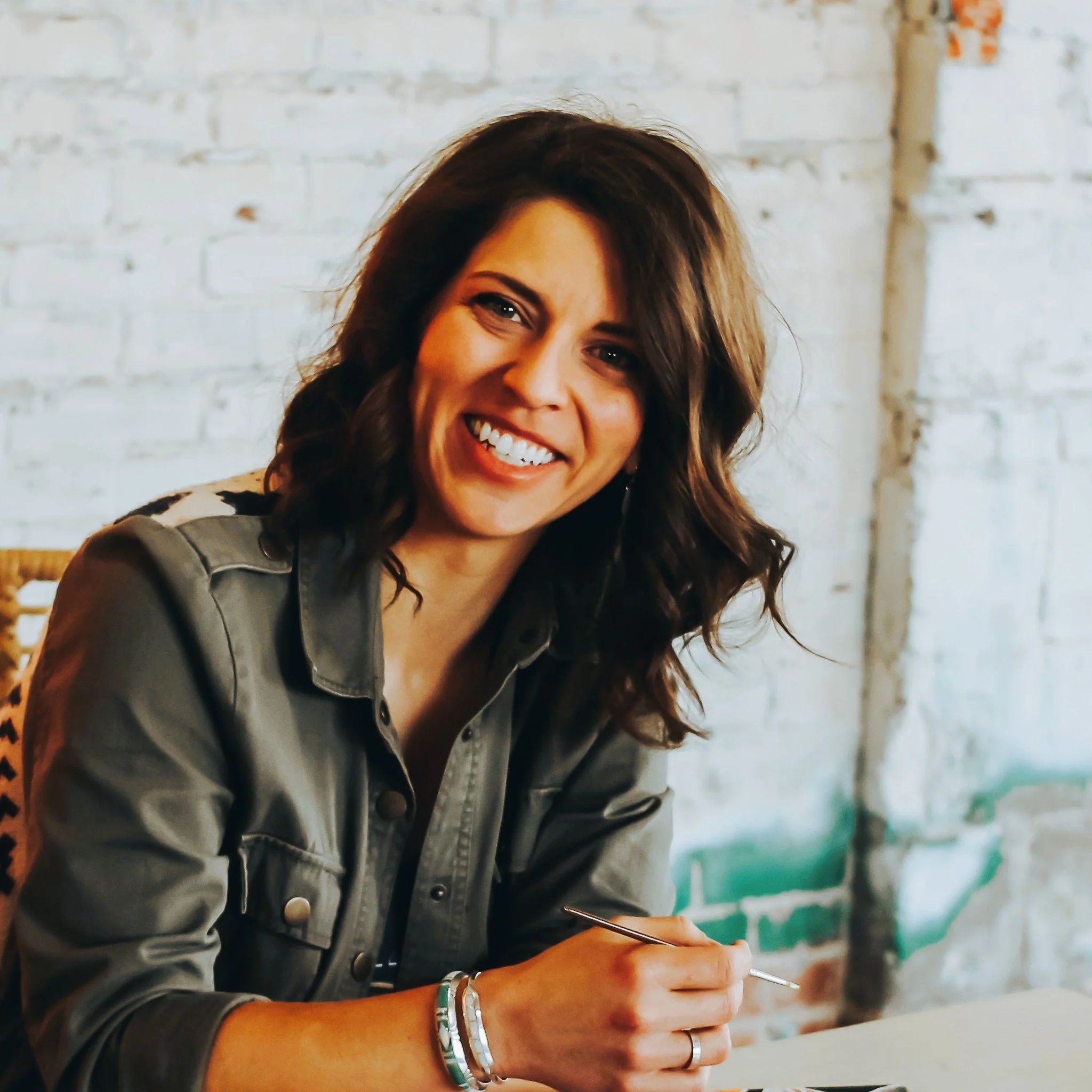 Artist Katie Hoffmeier in green jacket sitting at desk holding a paintbrush
