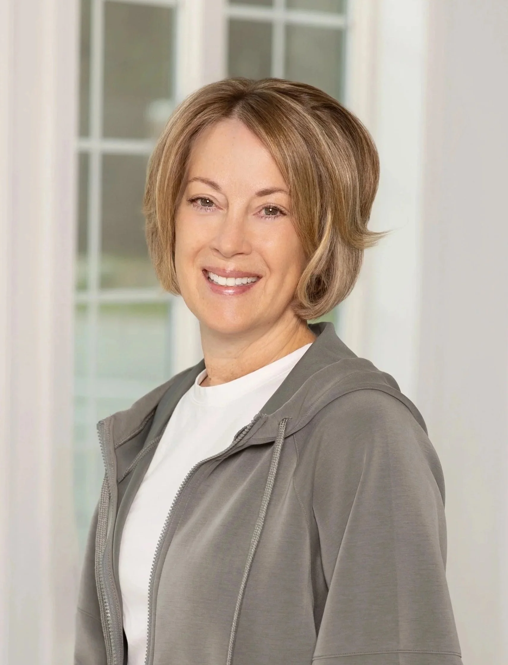 Photo of Deirdre Wenzel, yoga therapist, smiling, with short, light brown hair, wearing a white shirt and gray zip-up hoodie, standing indoors near windows.