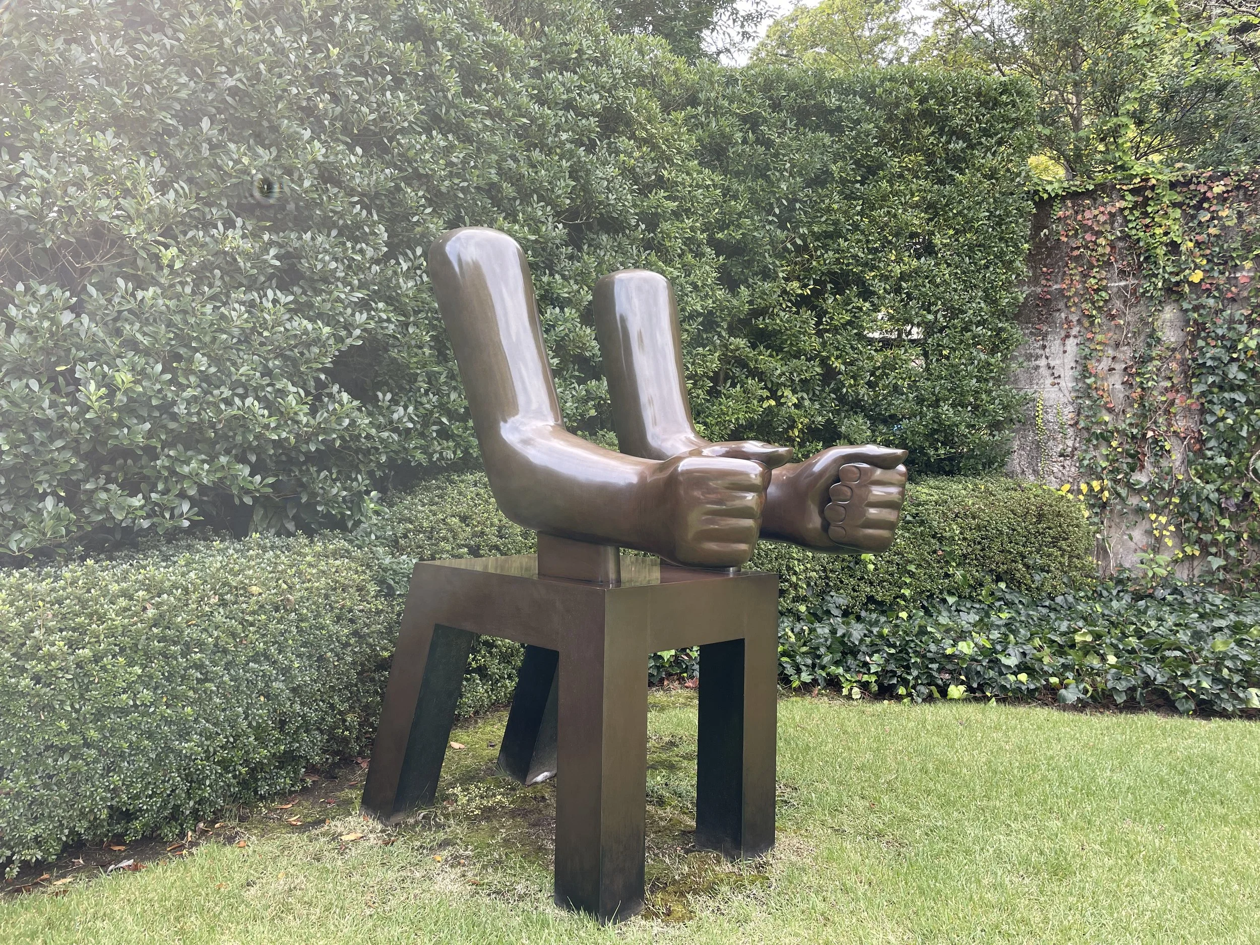 A bronze sculpture of two chairs with human fists as the seat and the backs as the armrests, situated outdoors on a grassy area with bushes and trees in the background.