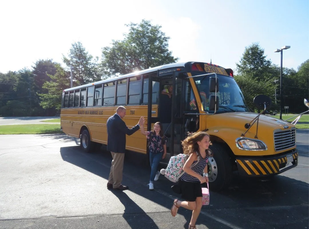 A school principal high-fives elementary school students as they step off a yellow school bus.