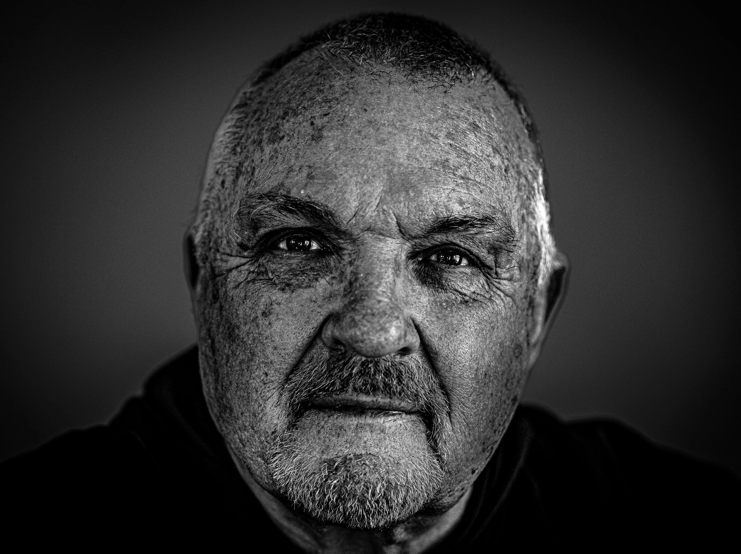 Close-up black and white portrait of an elderly man with a serious expression, gray hair and beard, looking directly at the camera.