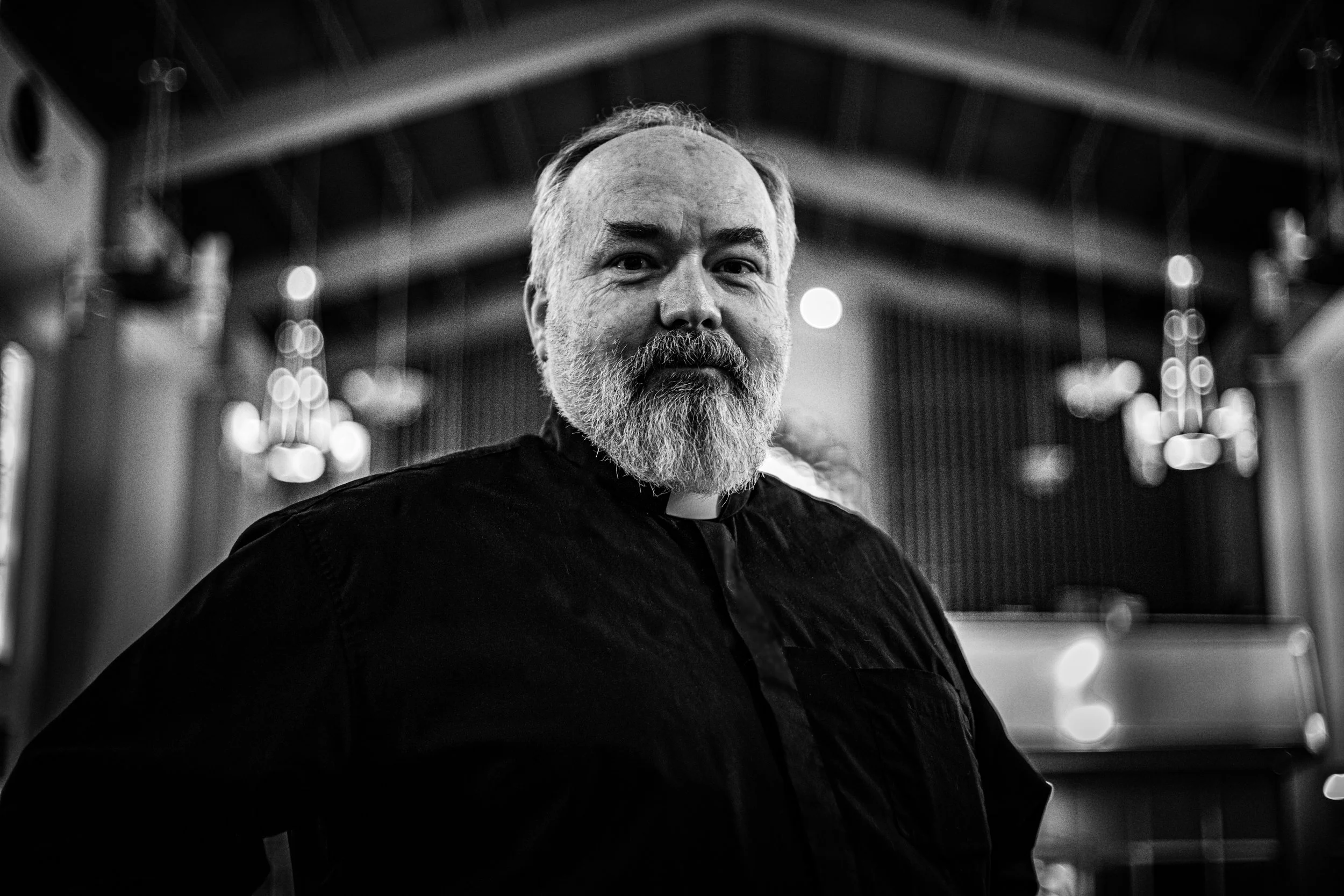 A bearded man in a dark shirt sitting in a room with ornate lighting fixtures and decorative wall panels.