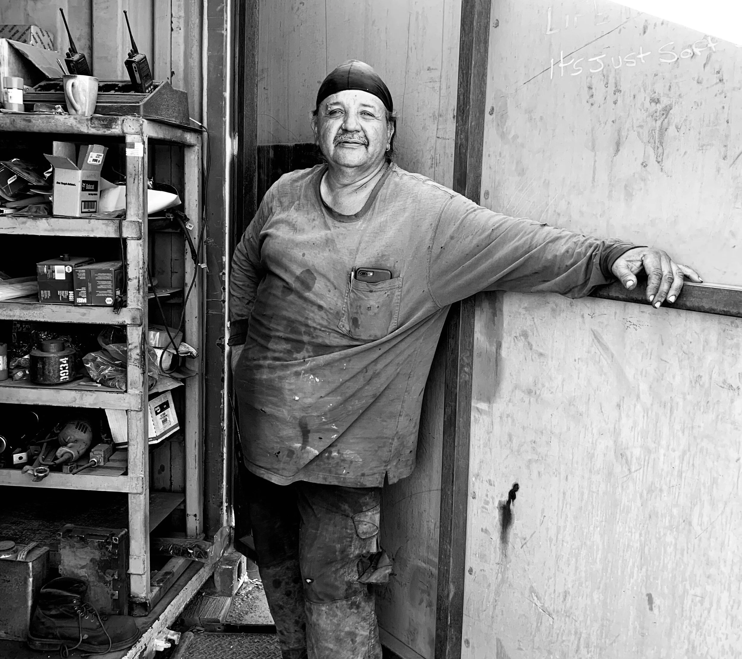 A man with a bandana and paint-stained clothing standing in a workshop, leaning against a wall with his arm outstretched, surrounded by tools and supplies.