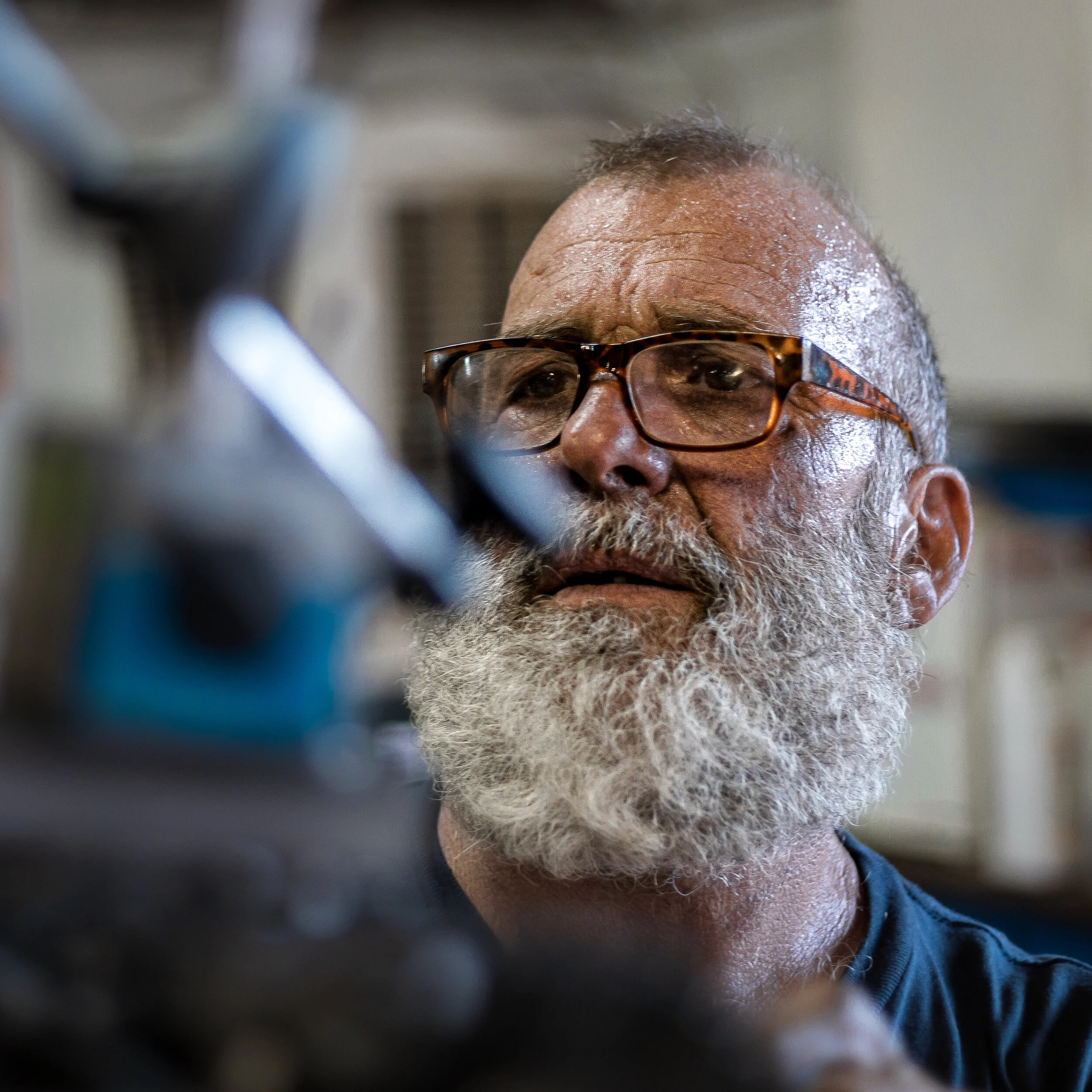 Close-up of an older man with glasses and a white beard working with machinery or tools indoors.