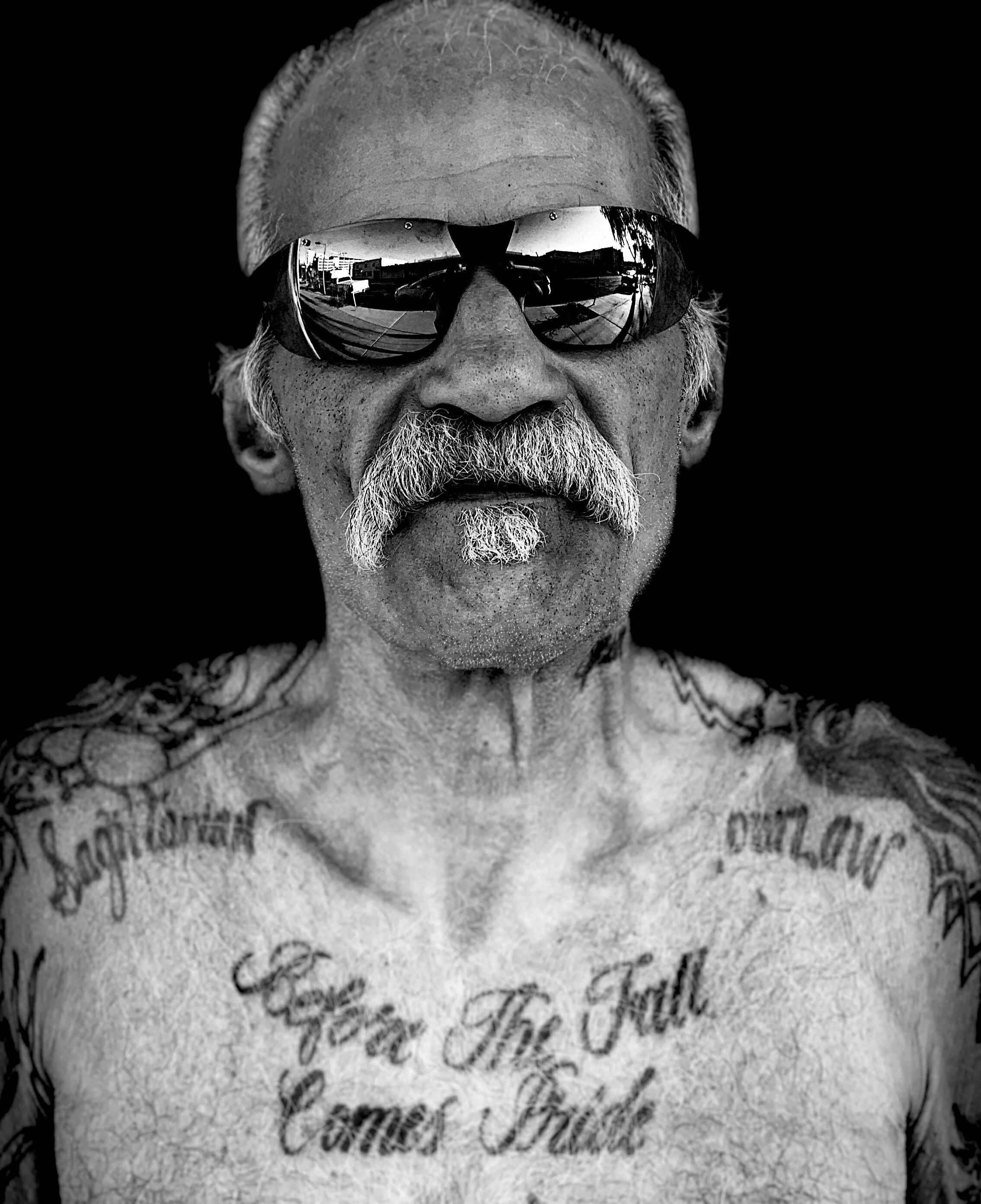 Black and white close-up photograph of an older man wearing sunglasses, with a large mustache and tattoos on his chest and shoulders.