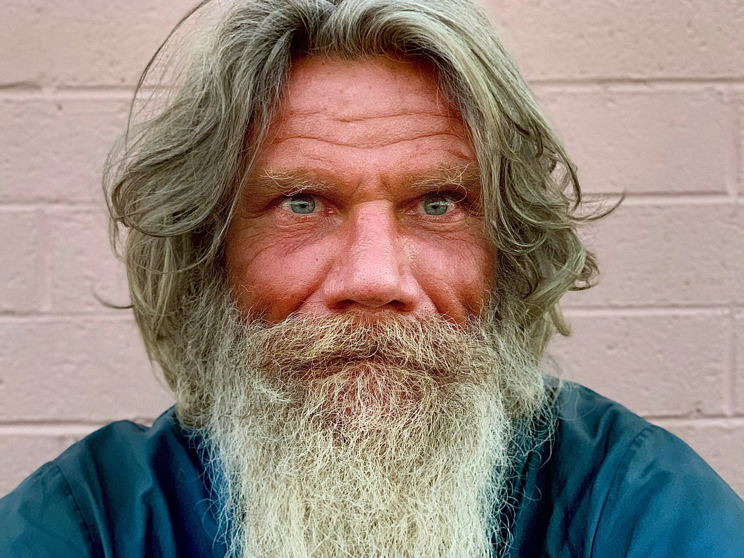 Close-up image of an older man with long gray hair, a full gray beard, and blue eyes, wearing a dark jacket against a plain beige brick wall.