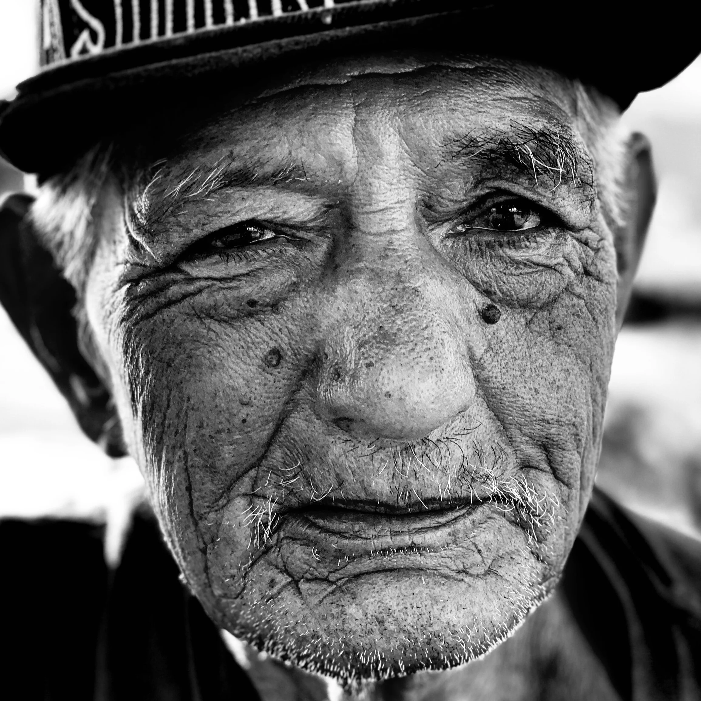 Close-up black-and-white portrait of an elderly man with deep wrinkles, wearing a hat.