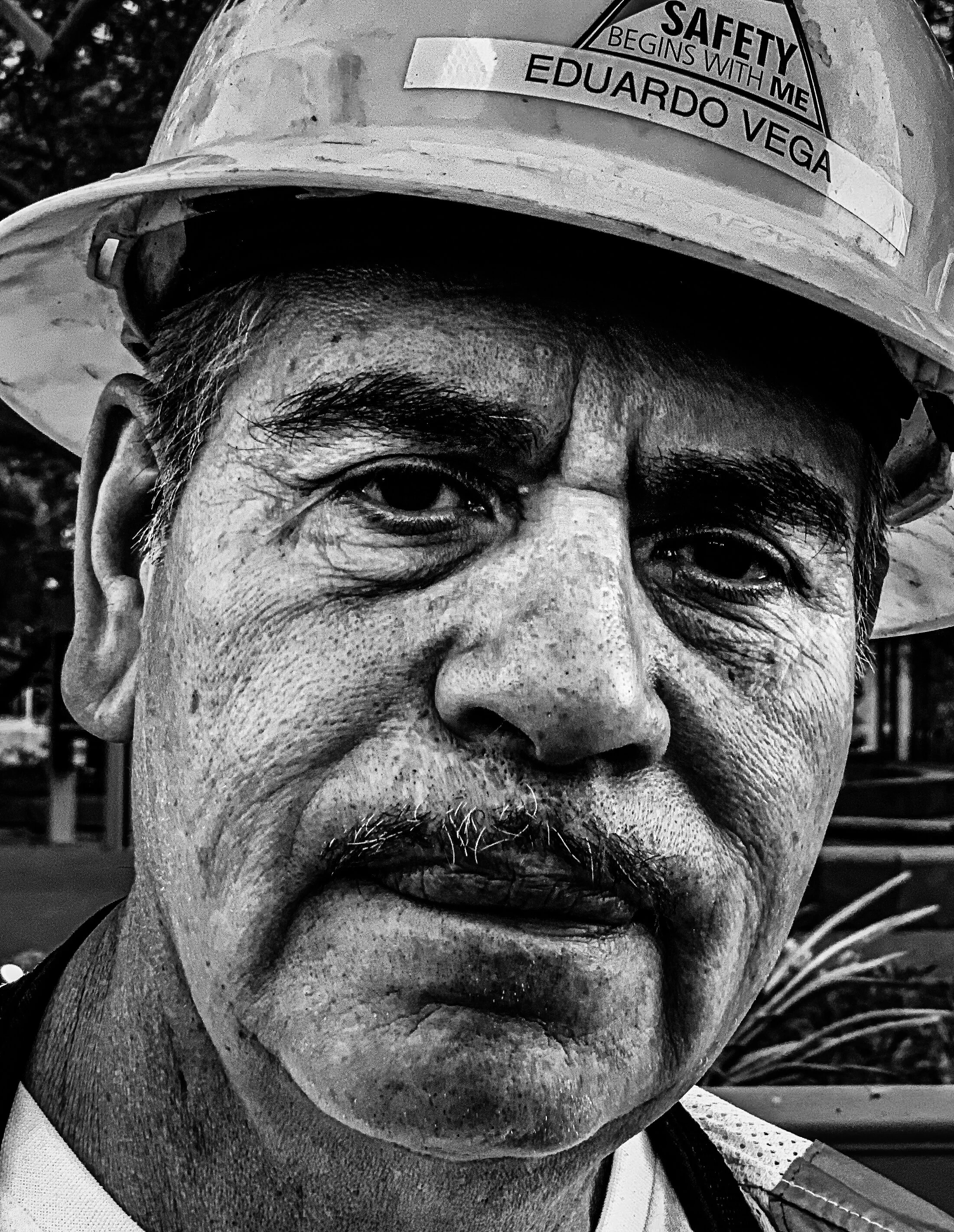 Close-up black and white photo of an older man wearing a construction helmet with a sticker that says "Safety begins with me Eduardo Vega."