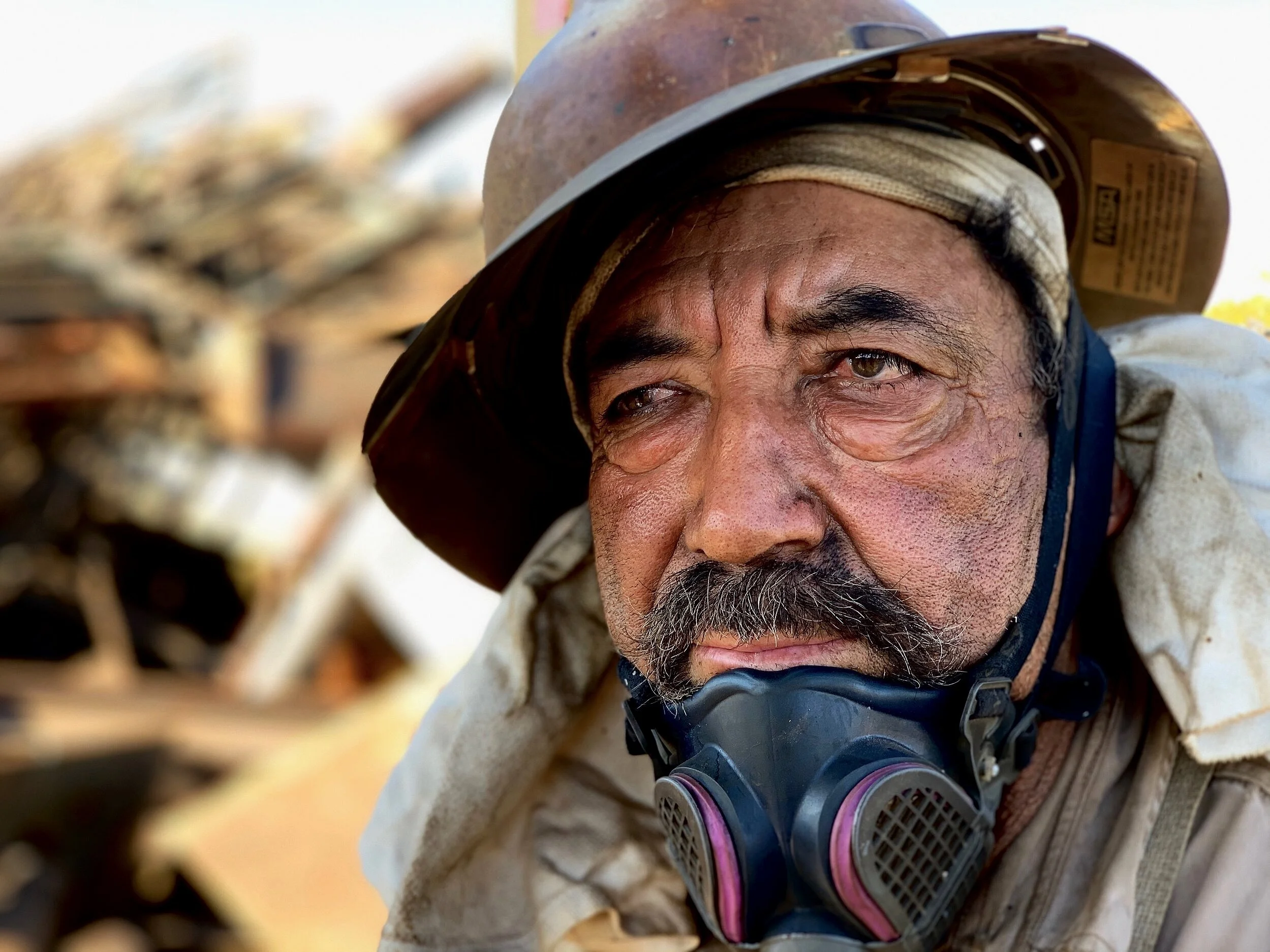 Close-up of a man wearing a safety helmet and a face mask around his neck, with a damaged industrial or construction site background.