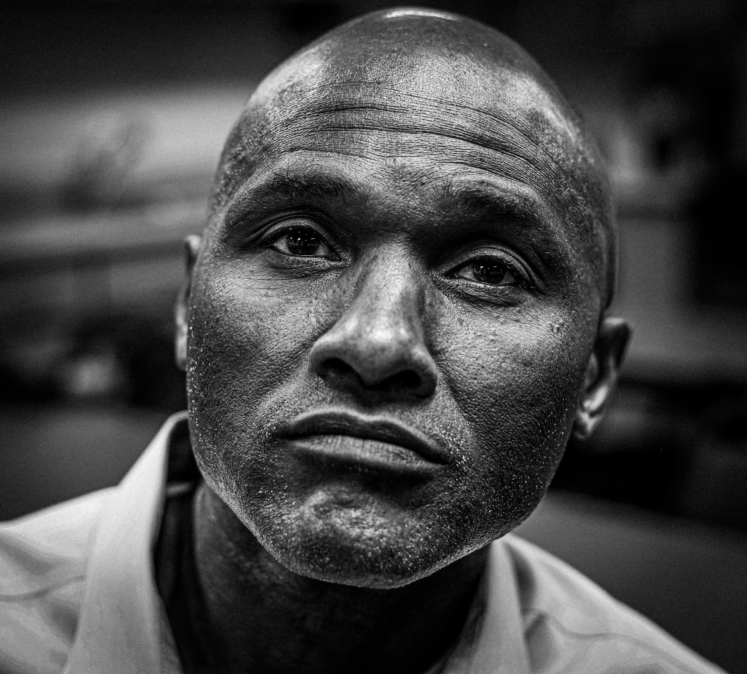A close-up black and white portrait of a man with a serious expression, visible pores, and a shaved head, wearing a collared shirt.
