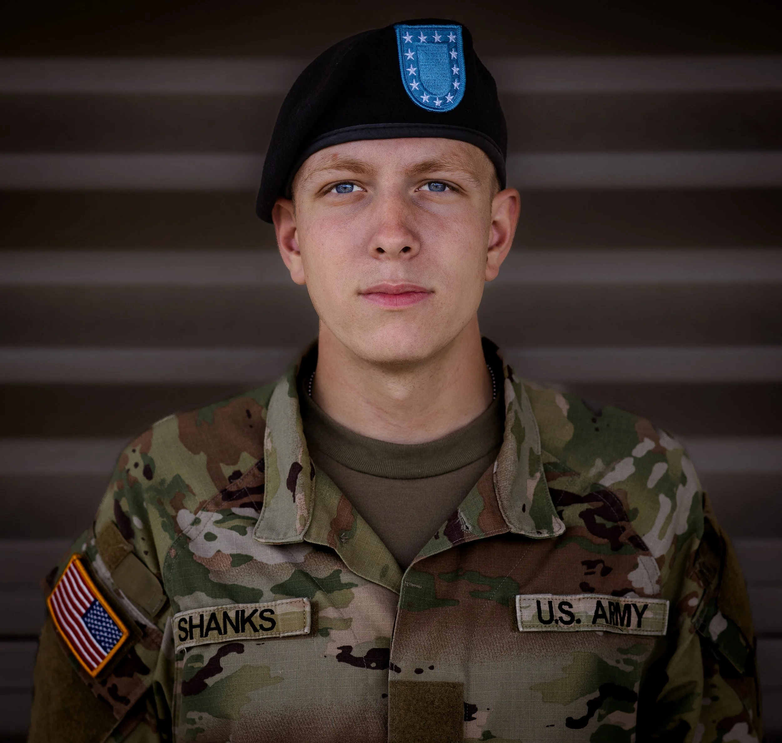 A young soldier in a U.S. Army camouflage uniform with a black beret, standing in front of a metal background. The uniform has name tag "SHANKS" and U.S. Army patch. The soldier has blue eyes and a neutral expression.