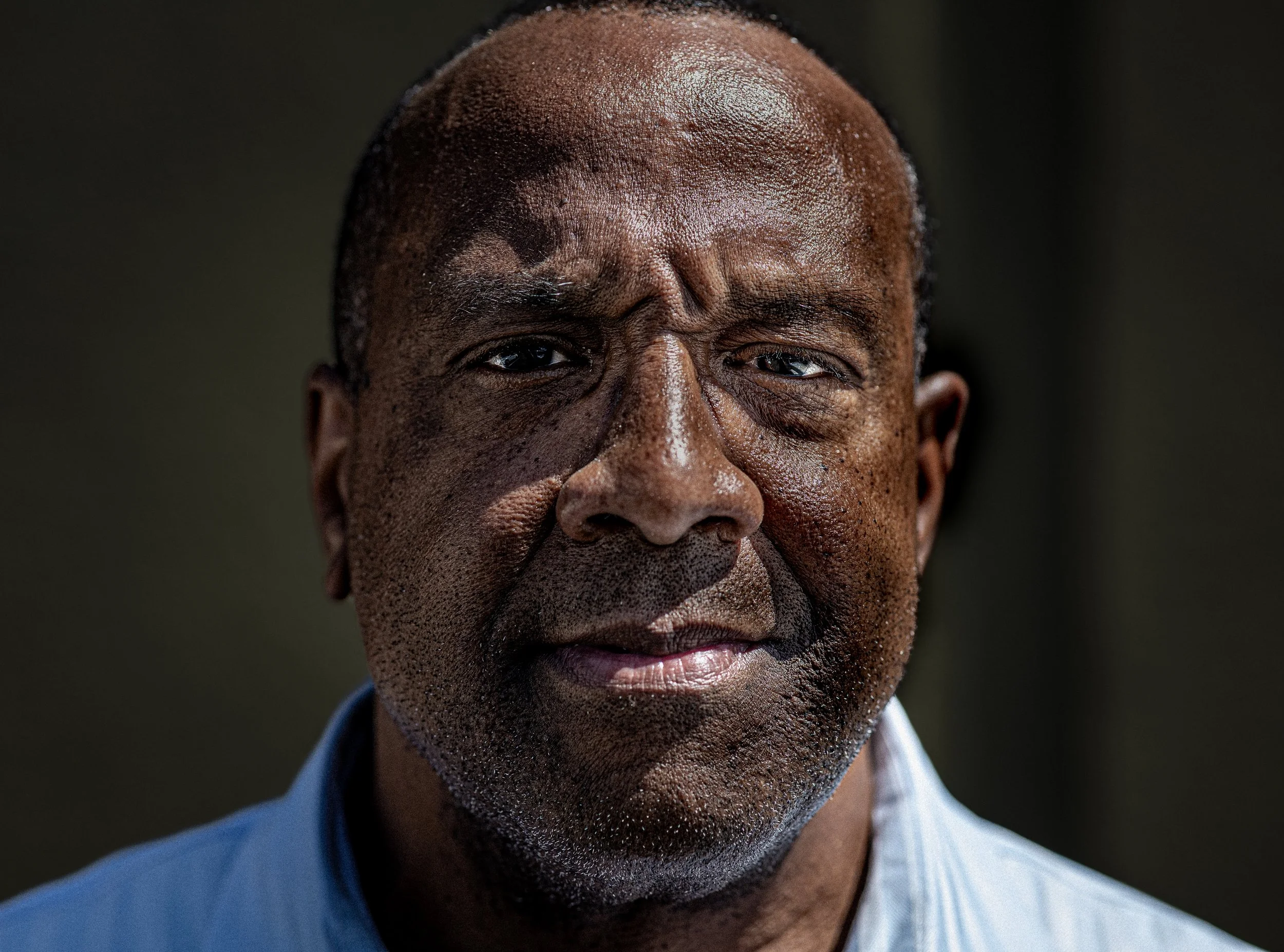 Close-up of a middle-aged man with dark skin, graying hair, and a thoughtful expression, wearing a blue collared shirt.