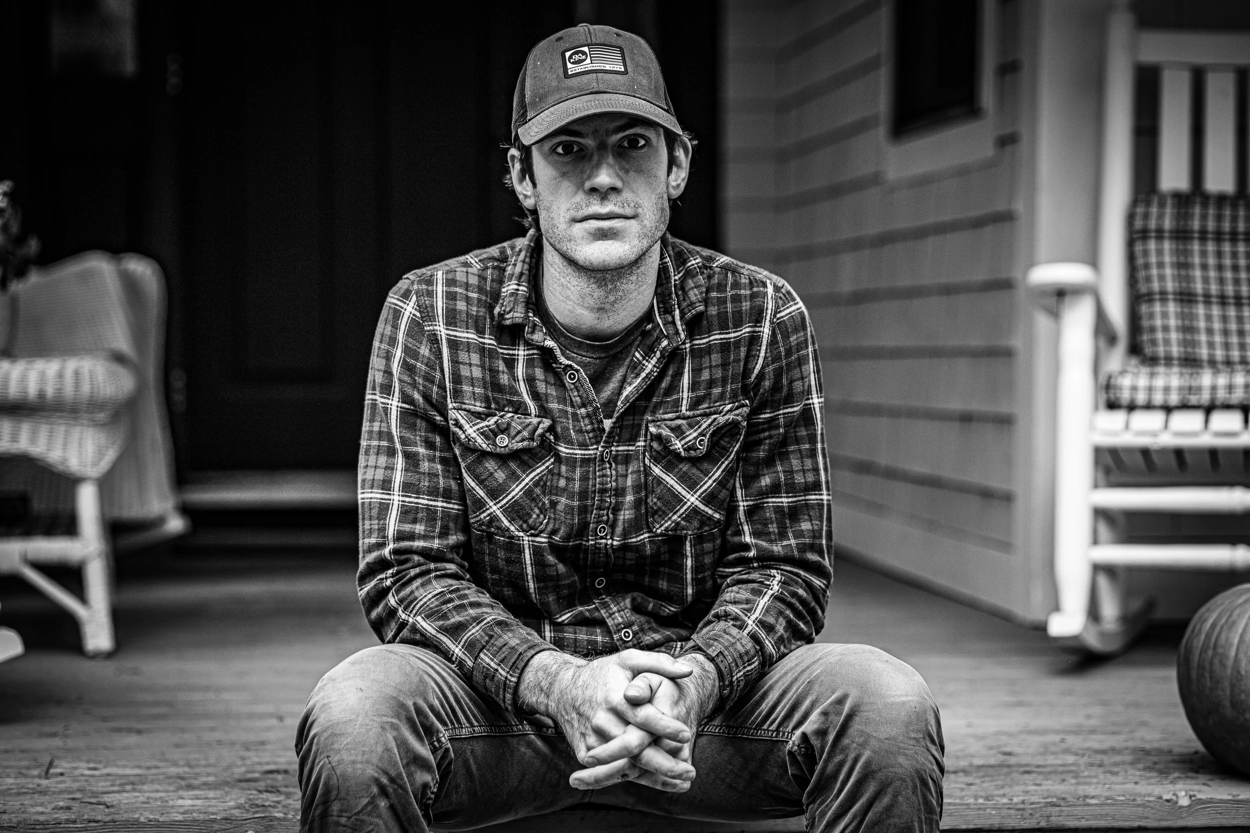A young man wearing a baseball cap, plaid shirt, and jeans sitting cross-legged on a wooden porch with his hands clasped. In the background, there are chairs and a pumpkin on the porch.