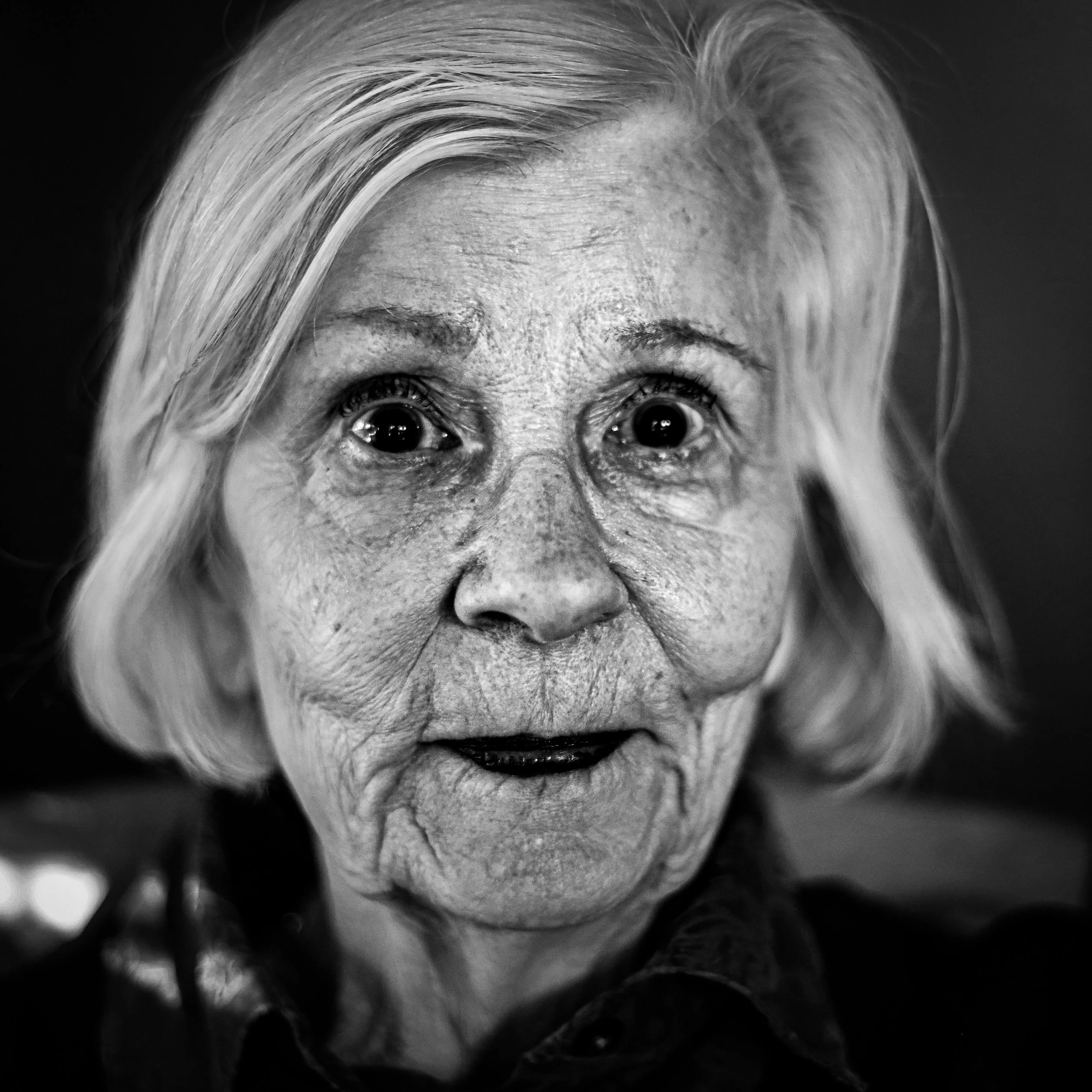 Close-up black and white photo of an elderly woman with short, light-colored hair, showing detailed facial wrinkles and a surprised or curious expression.