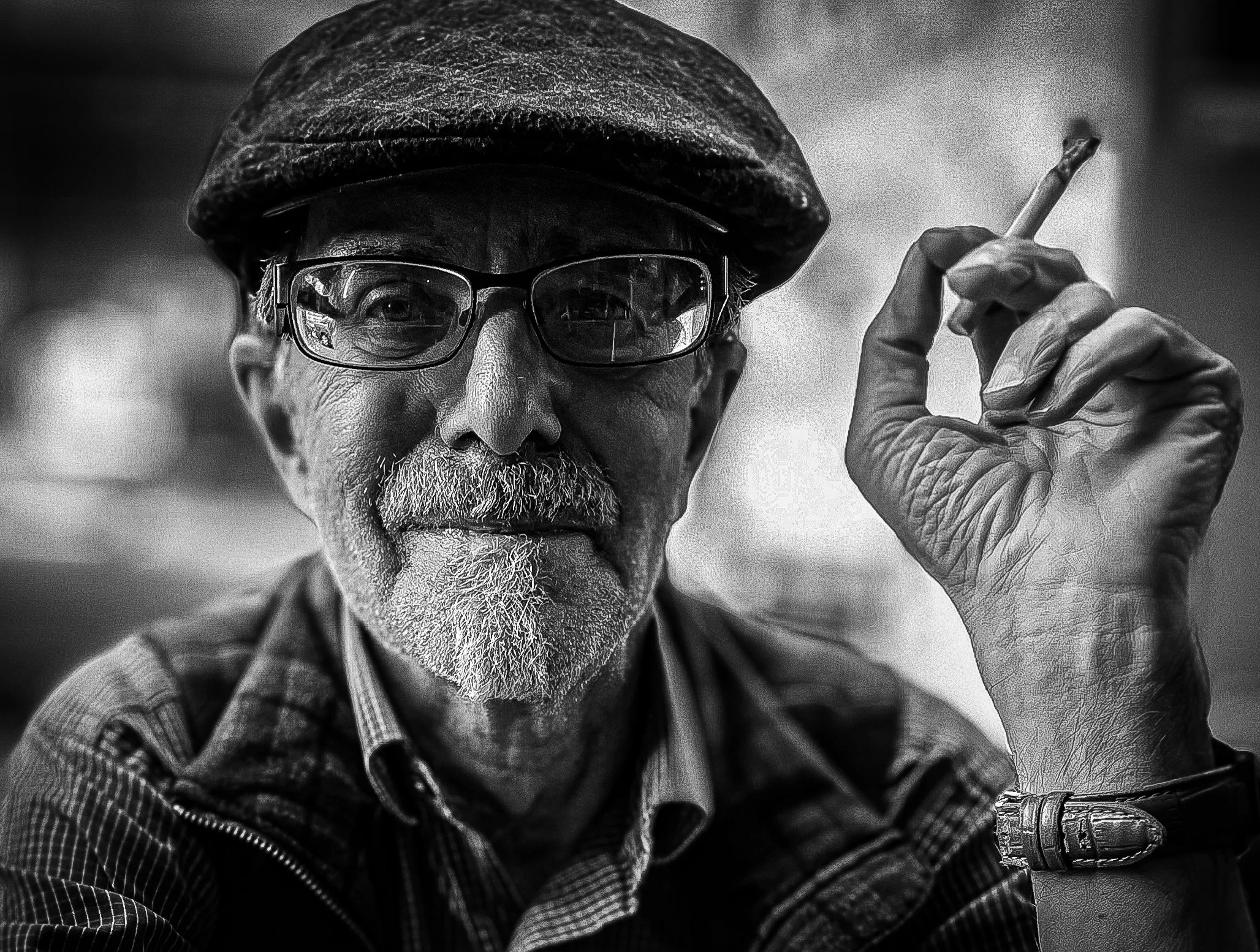 A black and white close-up of an elderly man wearing glasses, a cap, and a checkered shirt, holding a cigarette in his right hand with a wristwatch, looking directly at the camera.