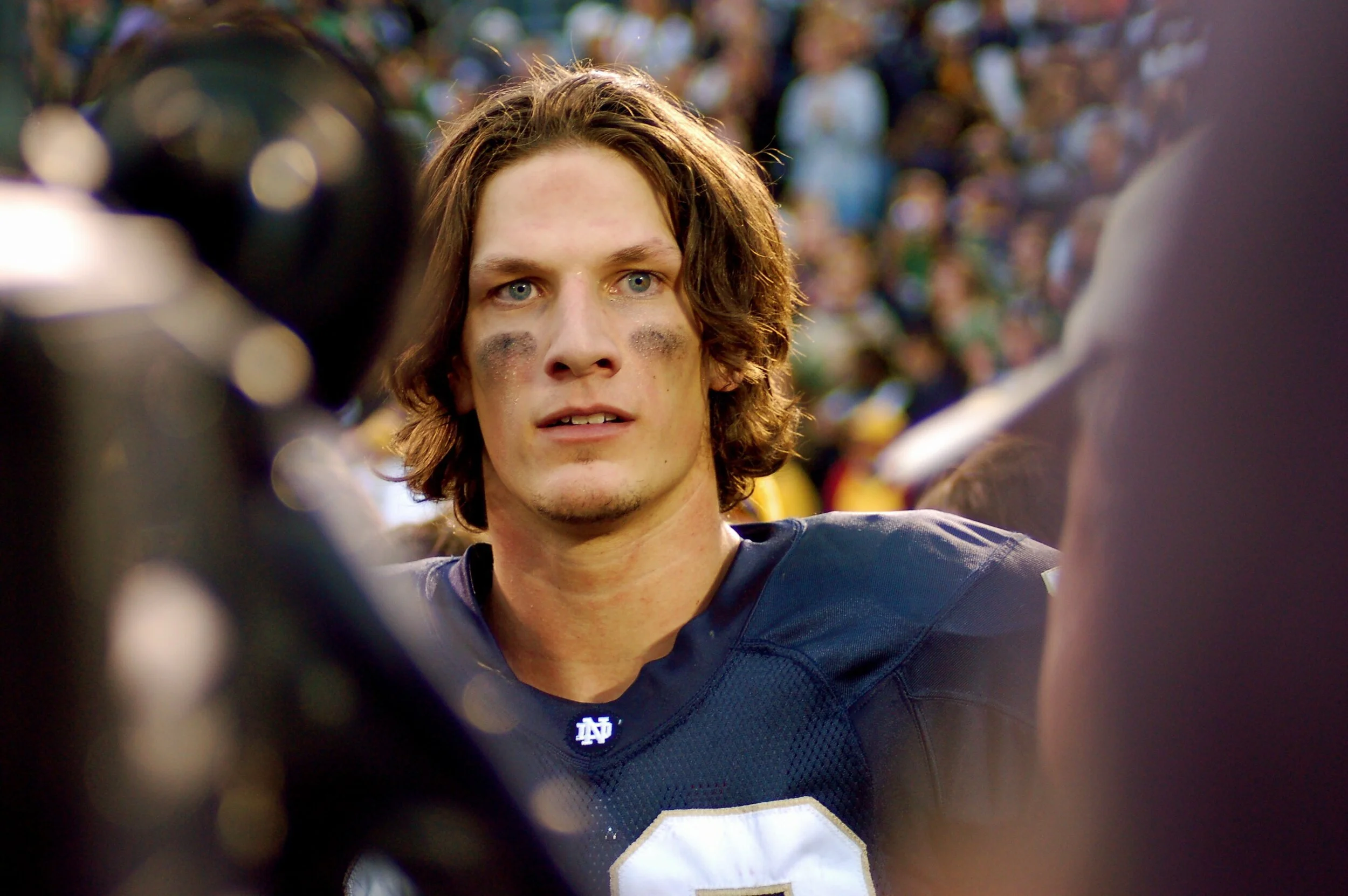 A football player with long brown hair and eye black is being interviewed on the field after a game, with a crowd in the background.
