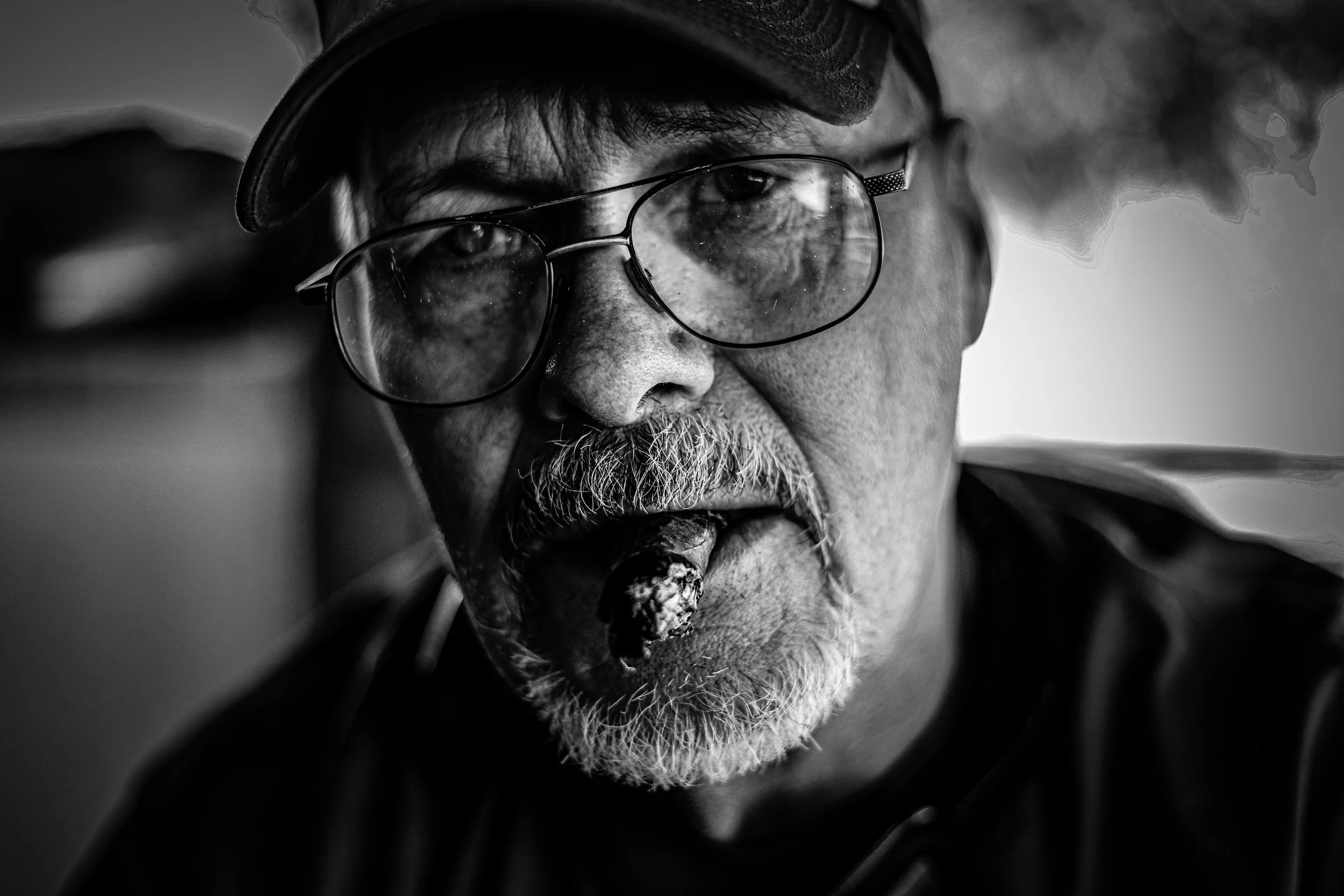 Close-up black and white photograph of an elderly man with glasses and a beard, smoking a pipe.