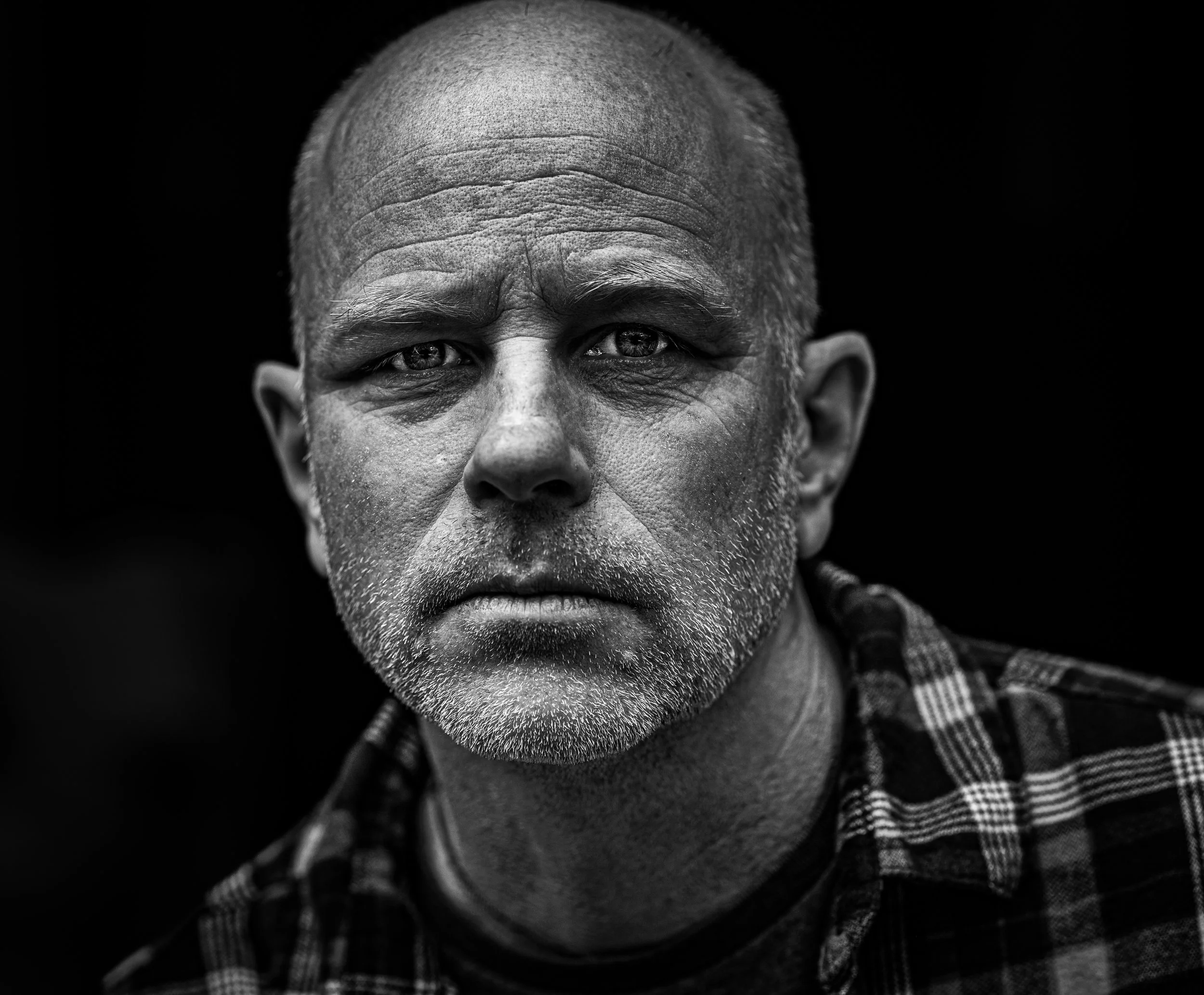 A black and white close-up portrait of a middle-aged man with a serious expression, short hair, a beard, and wearing a plaid shirt against a dark background.