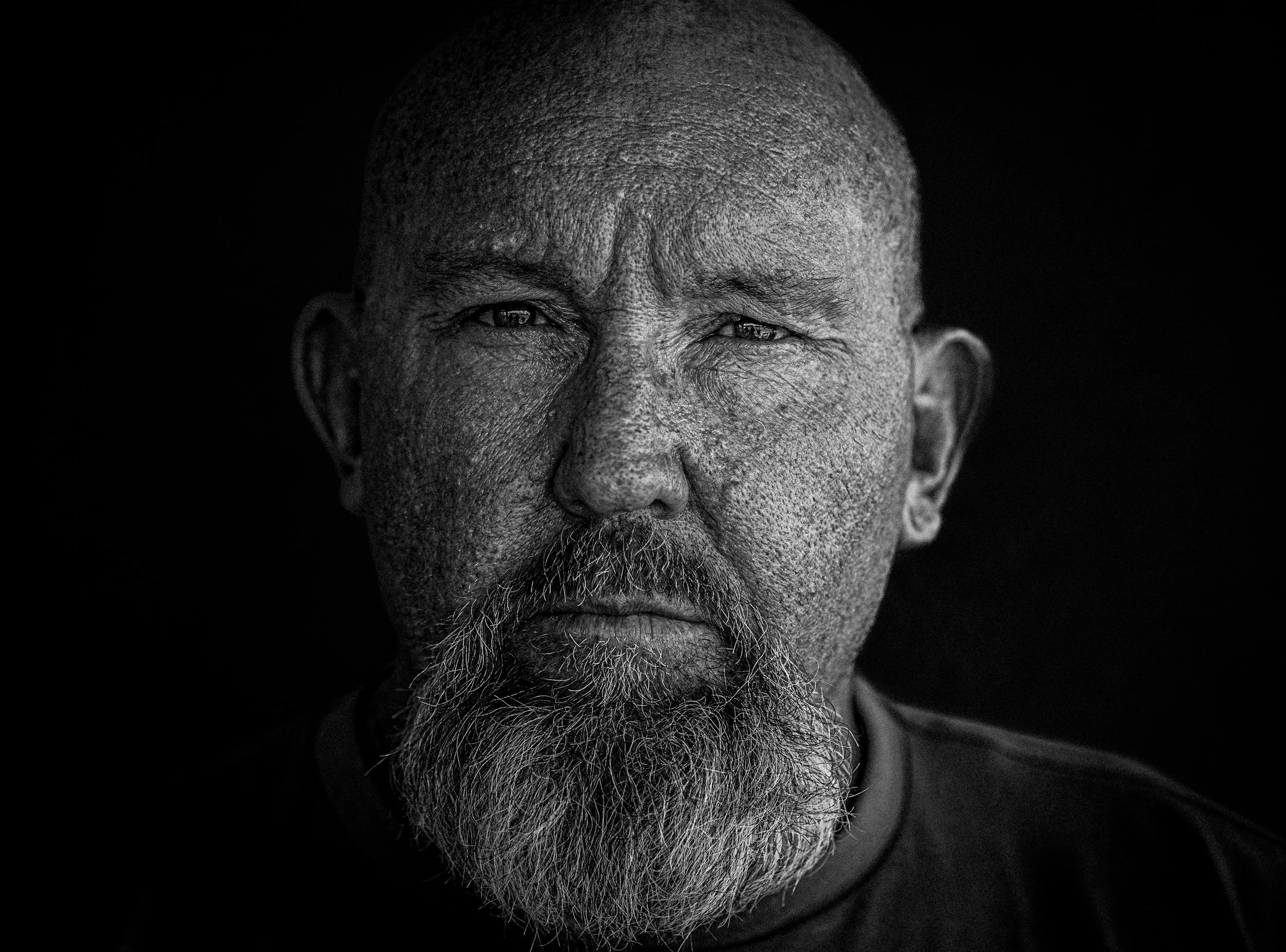 Black and white close-up portrait of an elderly man with a beard, looking directly at the camera against a dark background.