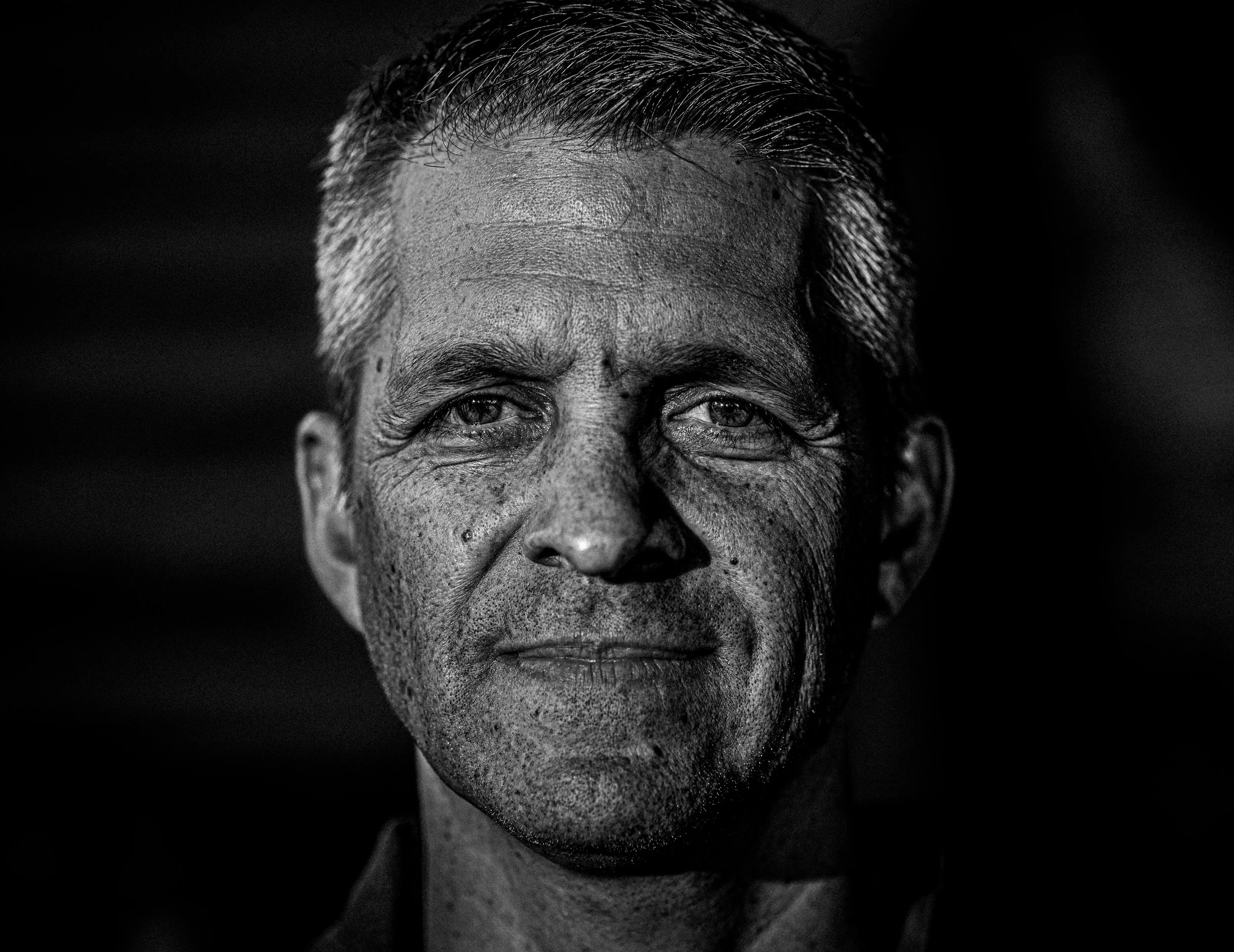 Black and white close-up portrait of an older man with gray hair and visible wrinkles, looking directly at the camera.