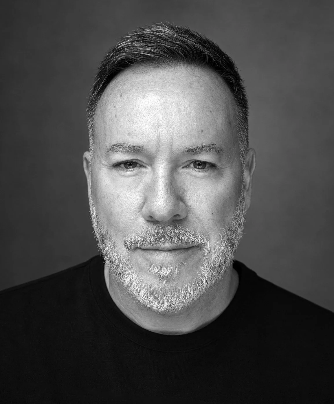 Black and white portrait of a middle-aged man with short hair, a beard, and a confident expression, wearing a black shirt.