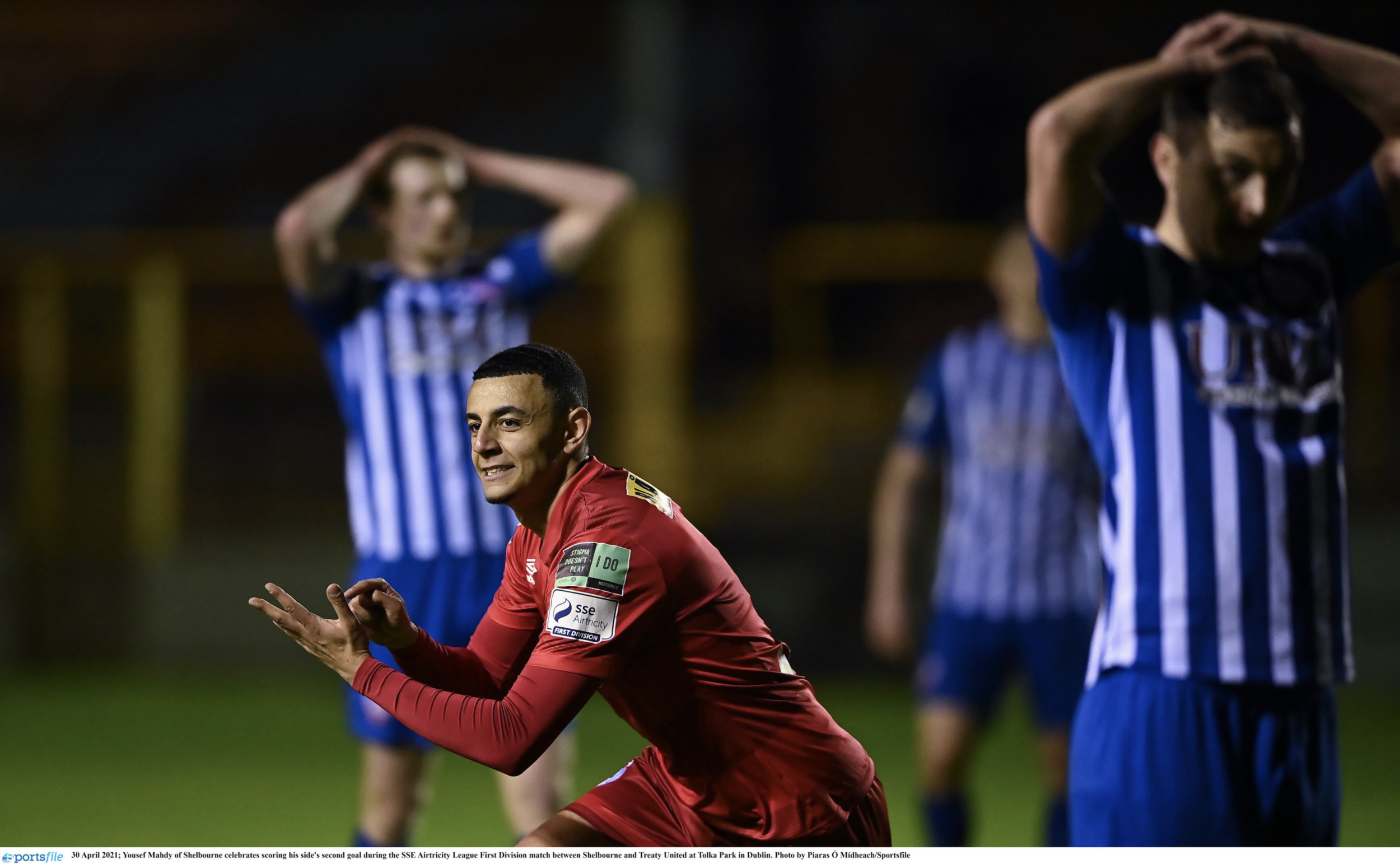 Soccer player in red celebrating a goal on the field at night, with other players in blue and white striped jerseys in the background.