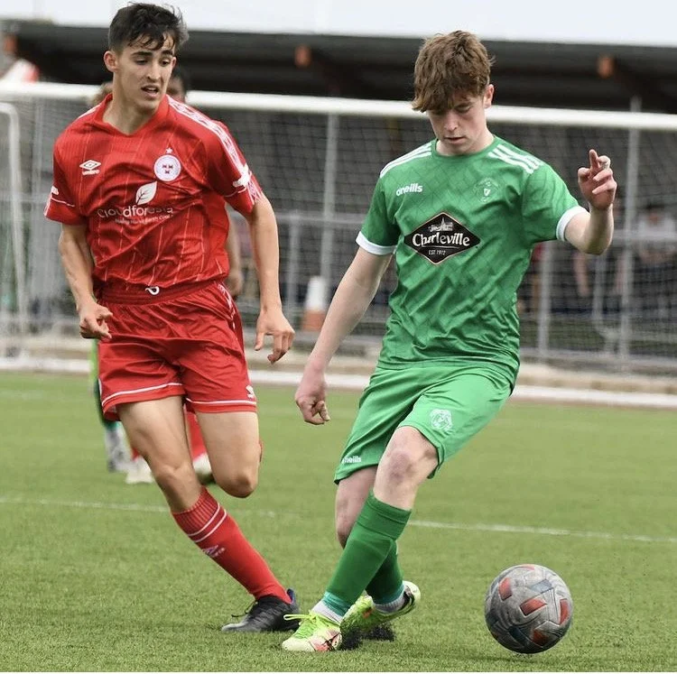 Two soccer players competing for the ball on the field, one in a red uniform and the other in a green uniform.